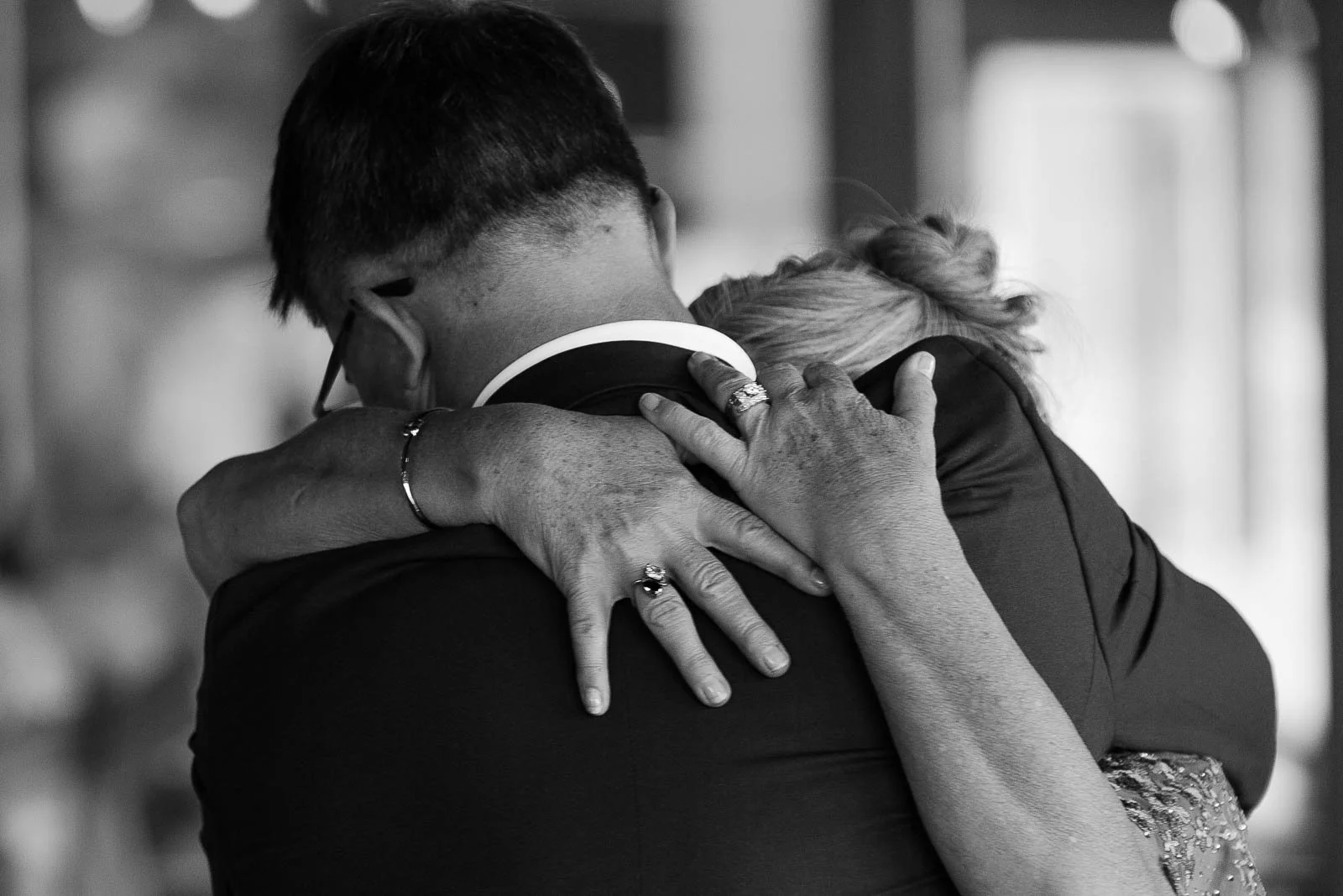  A close-up, black-and-white photo of two people embracing tightly, their faces hidden. One person wears a suit; the other's hand, featuring a ring, rests gently on the suit’s back, suggesting a moment of deep emotion. 