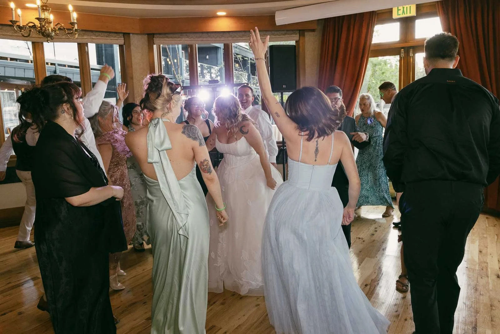  A group of people in formal attire dance and celebrate indoors at a well-lit event, possibly a wedding reception, with one woman raising her arm and others smiling around her. 