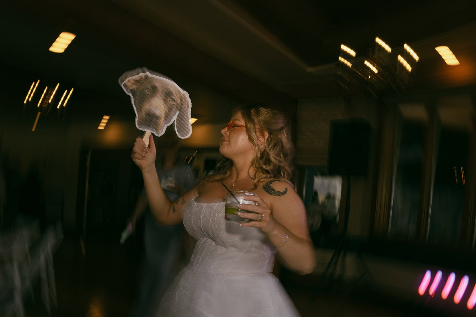  A woman in a white dress holds a drink and a cutout of a dog's face at a dimly lit indoor event, with blurred lights and motion effect in the background. 