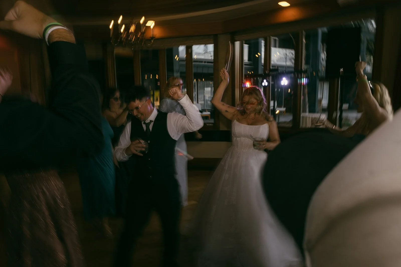  A bride and groom dance joyfully at their wedding reception, surrounded by guests. The scene is lively with blurred motion, warm lighting, and casual, celebratory atmosphere. 