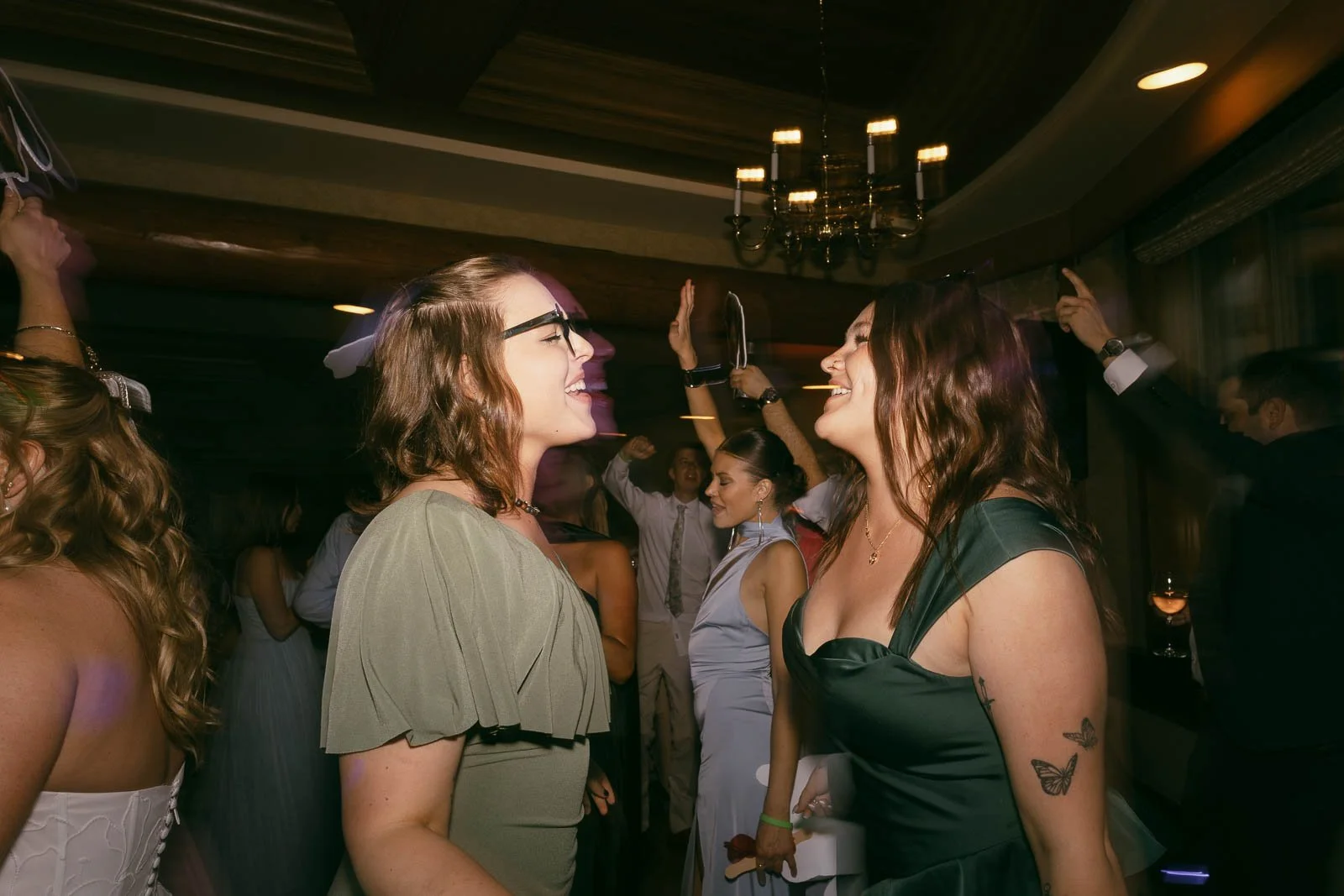  Two women in dresses smile and dance at a lively indoor party, surrounded by other guests. The room is warmly lit with a chandelier above and people holding their hands up in the background. 