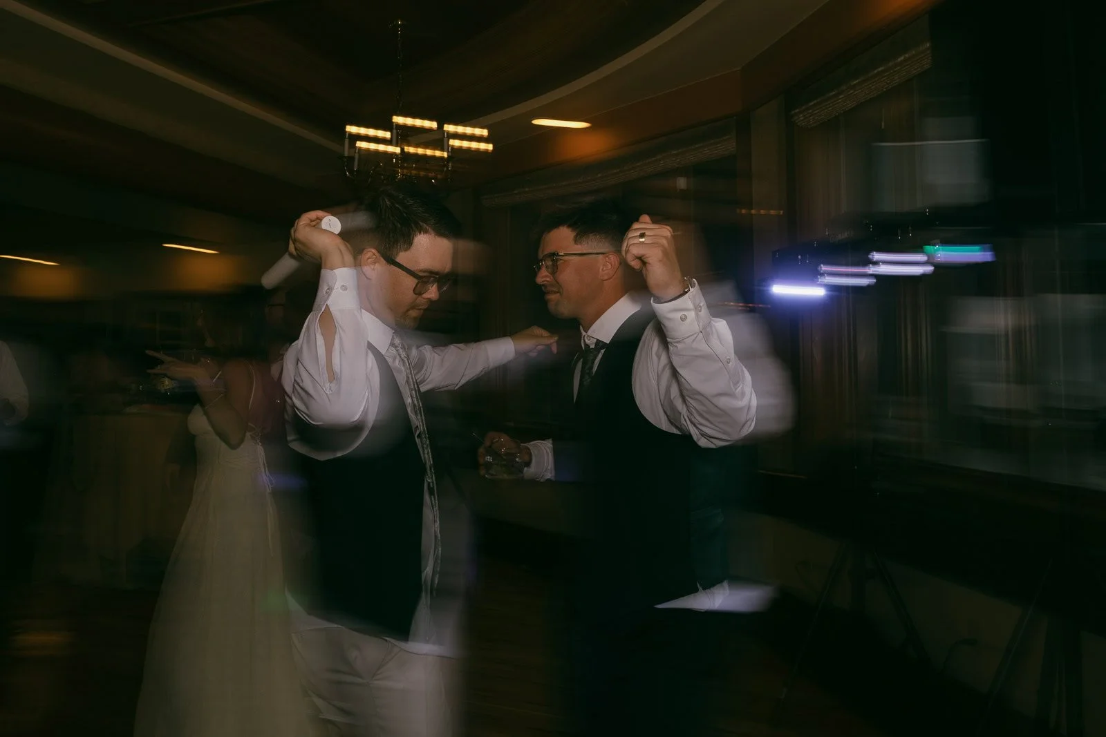  Two men in vests and dress shirts dance together at an indoor event, holding hands and smiling, with blurred lights and people in the background creating a lively atmosphere. 