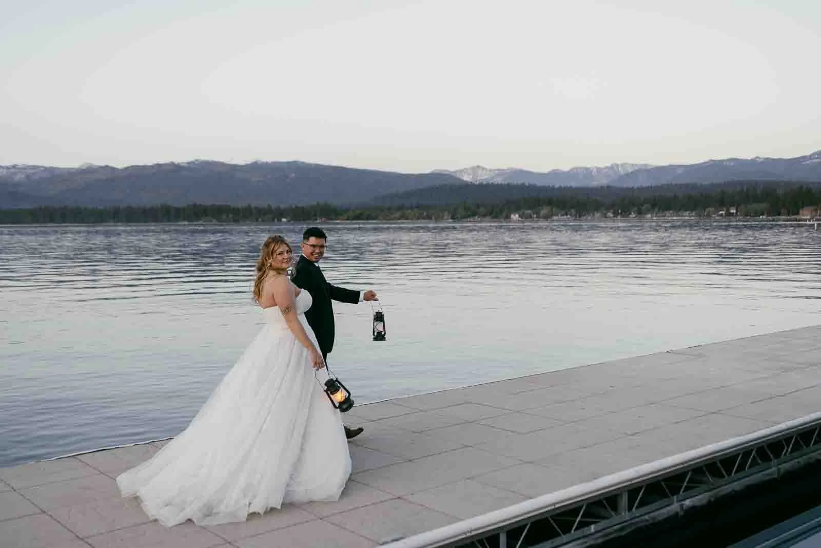 A smiling newlywed couple carries glowing lanterns as they stroll along a lakeside dock at sunset, mountains rising behind them.