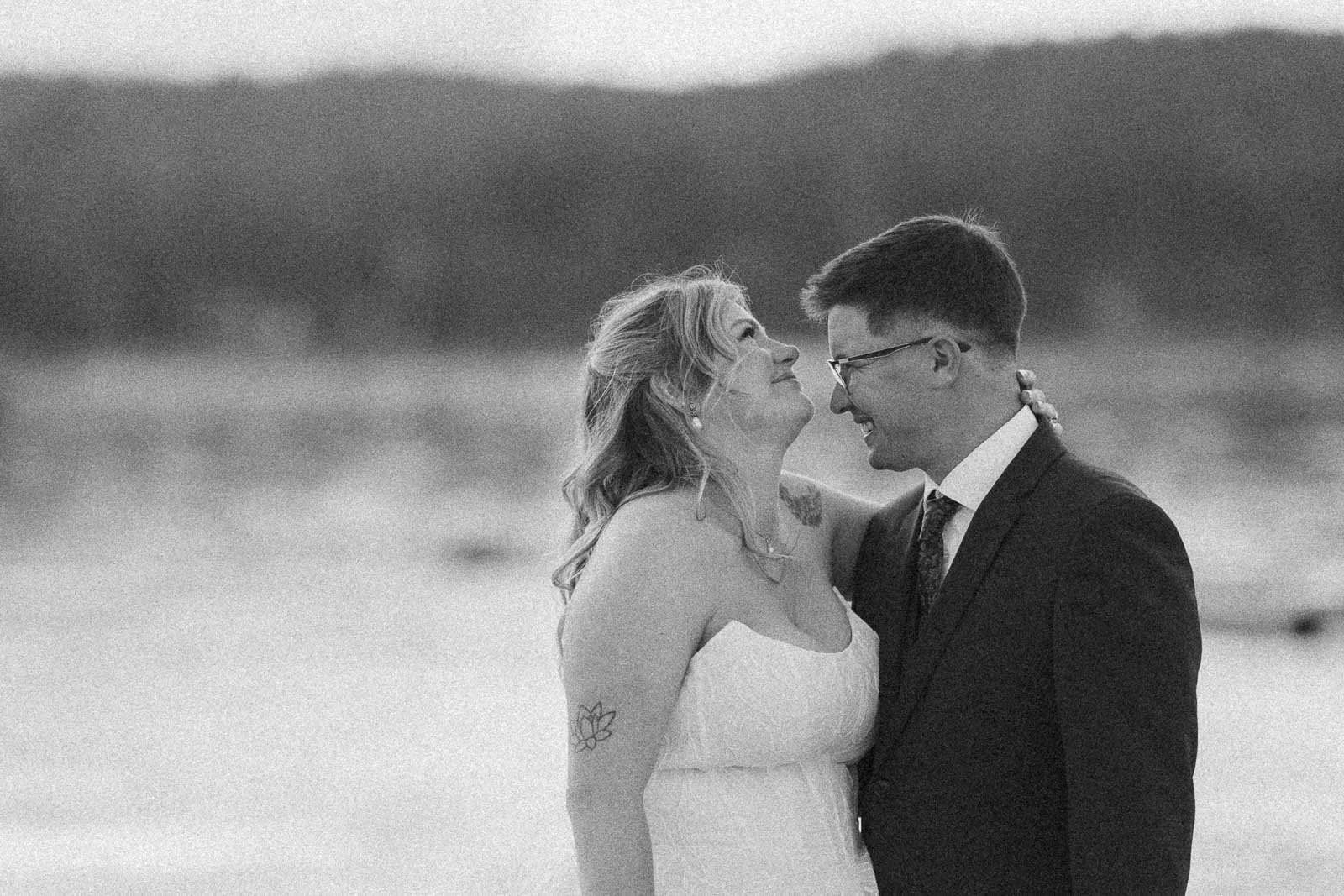 An outdoor wedding portrait of a couple gazing affectionately at one another, framed by soft, out-of-focus water and foliage.