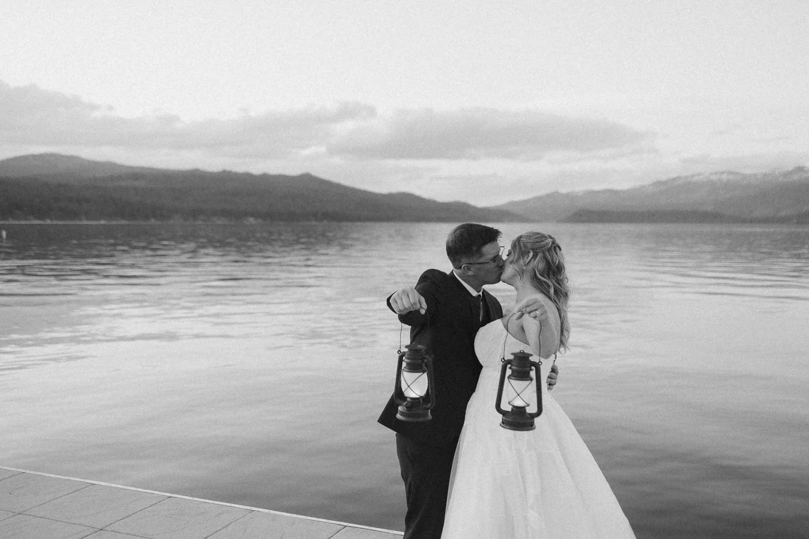  A bride and groom holding lanterns kiss by a calm lake, with mountains and clouds in the background. The photo is in black and white, creating a romantic, timeless atmosphere. 