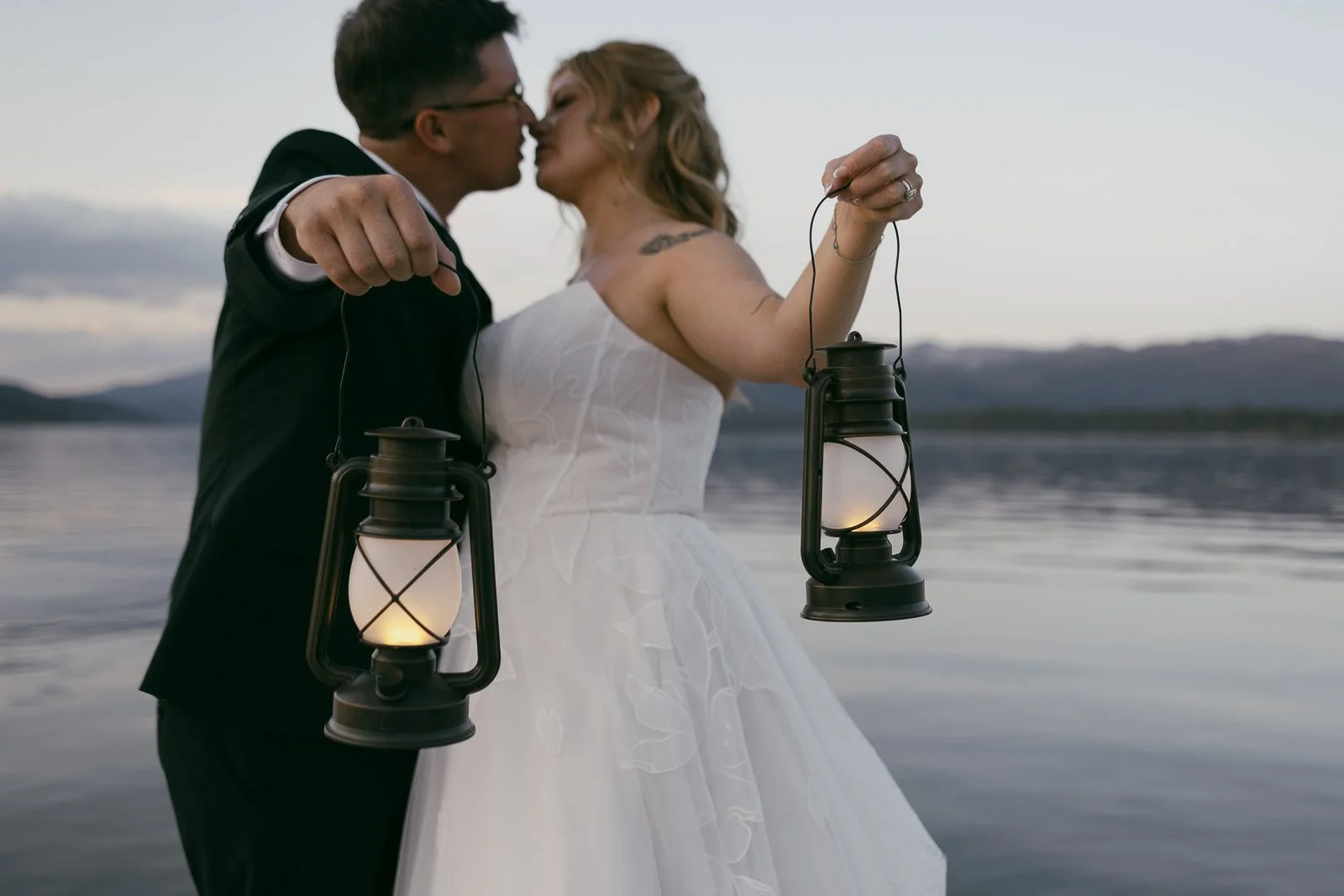  A bride and groom kiss while holding lit lanterns, standing by a calm lake with mountains in the background at dusk. 