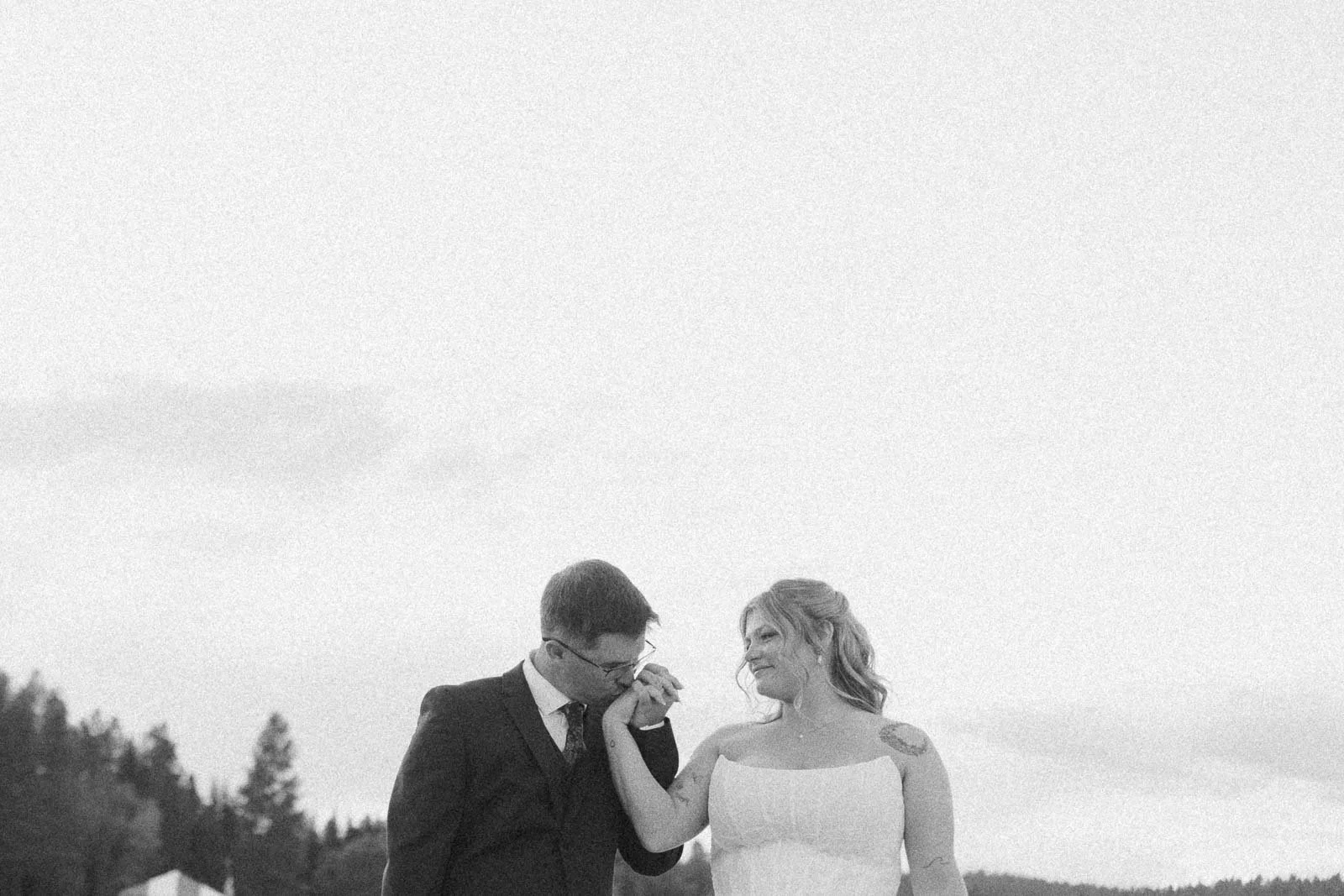  A man in a suit kisses the hand of a woman in a white dress outdoors. They smile at each other against a light sky, with trees and a building in the background. The image is in black and white. 