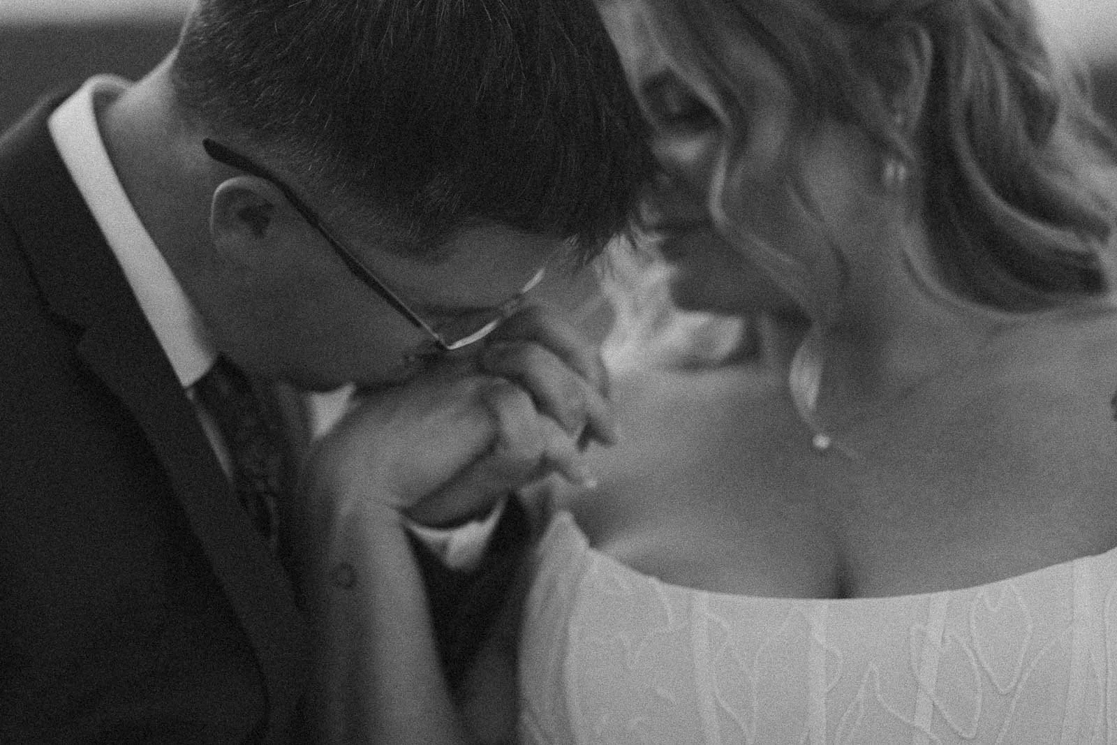  A groom gently kisses the bride’s hand as they stand close together. The black-and-white image captures an intimate, tender moment between the newlyweds, highlighting emotion and connection. 