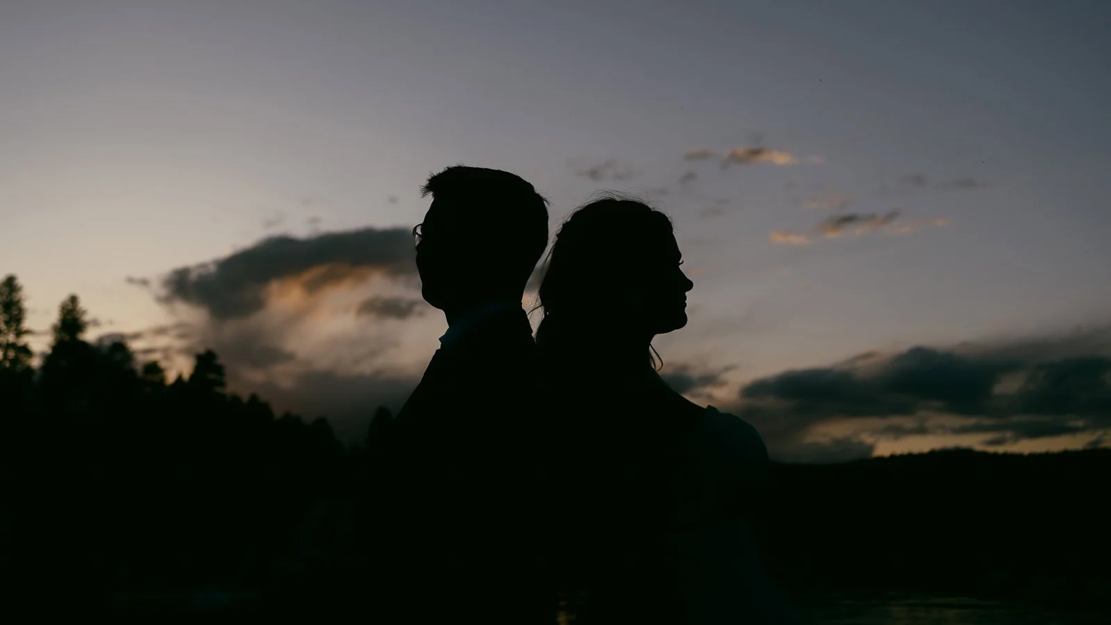  Silhouettes of two people standing back to back outdoors at sunset, with a dramatic sky and trees in the background. 