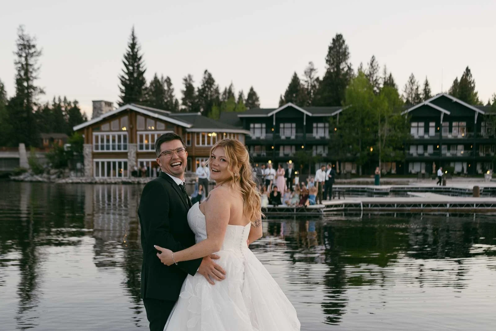 A bride and groom smile at the camera while standing beside a lake, with modern lodge buildings and wedding guests visible in the background. 