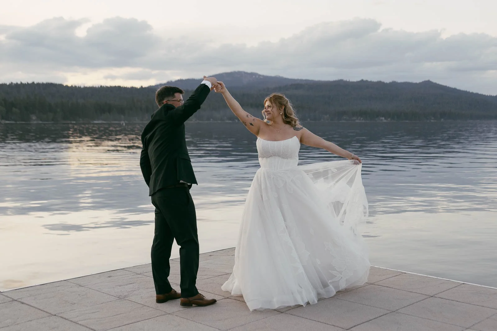  A gown-clad bride and her groom share a dance on a quiet lake dock, encircled by mountain peaks under a cloud-streaked sky. 