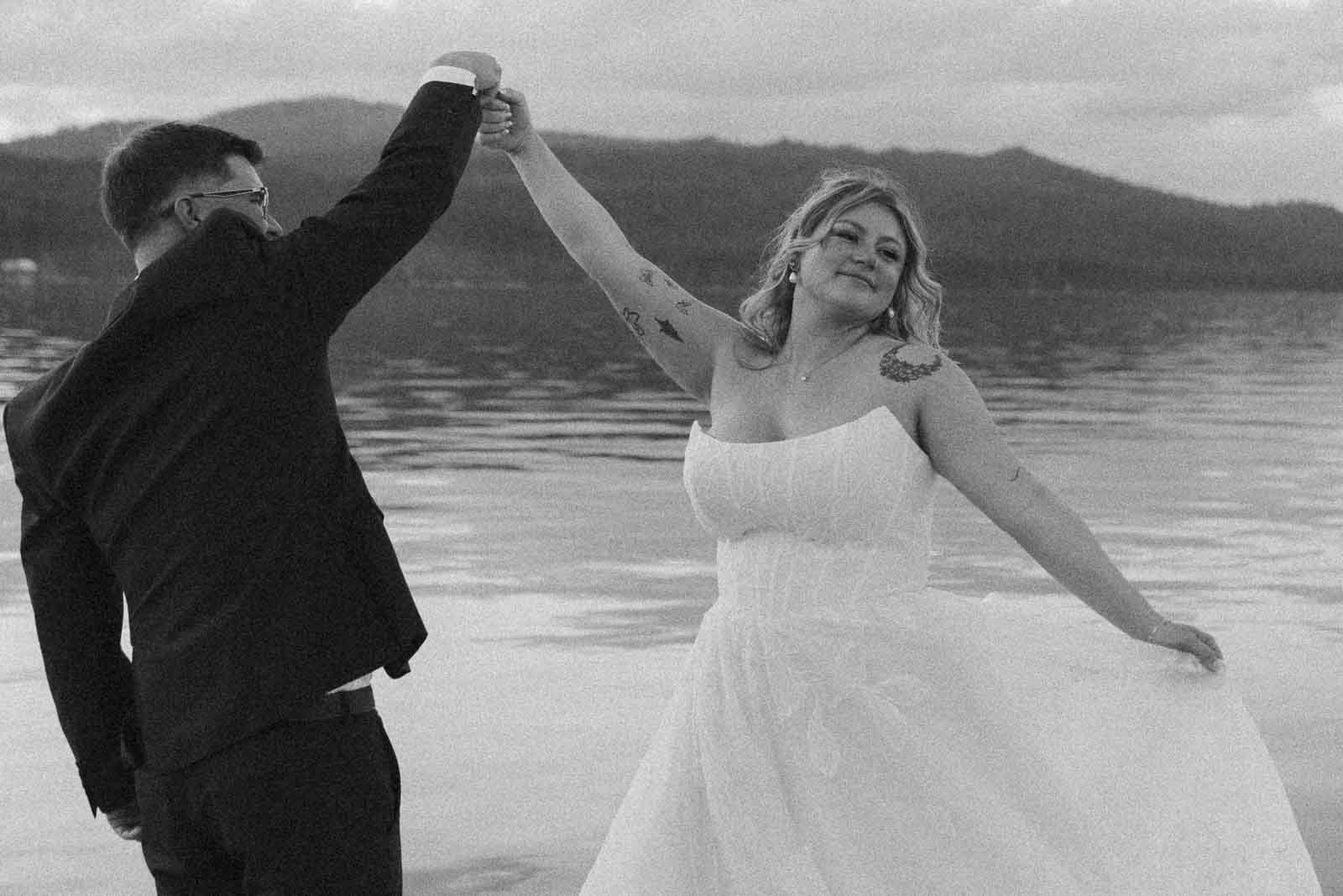  By a still lake, a bride twirls joyfully in her dress as the groom holds her hand mid-dance, mountains framing the scene beyond. 