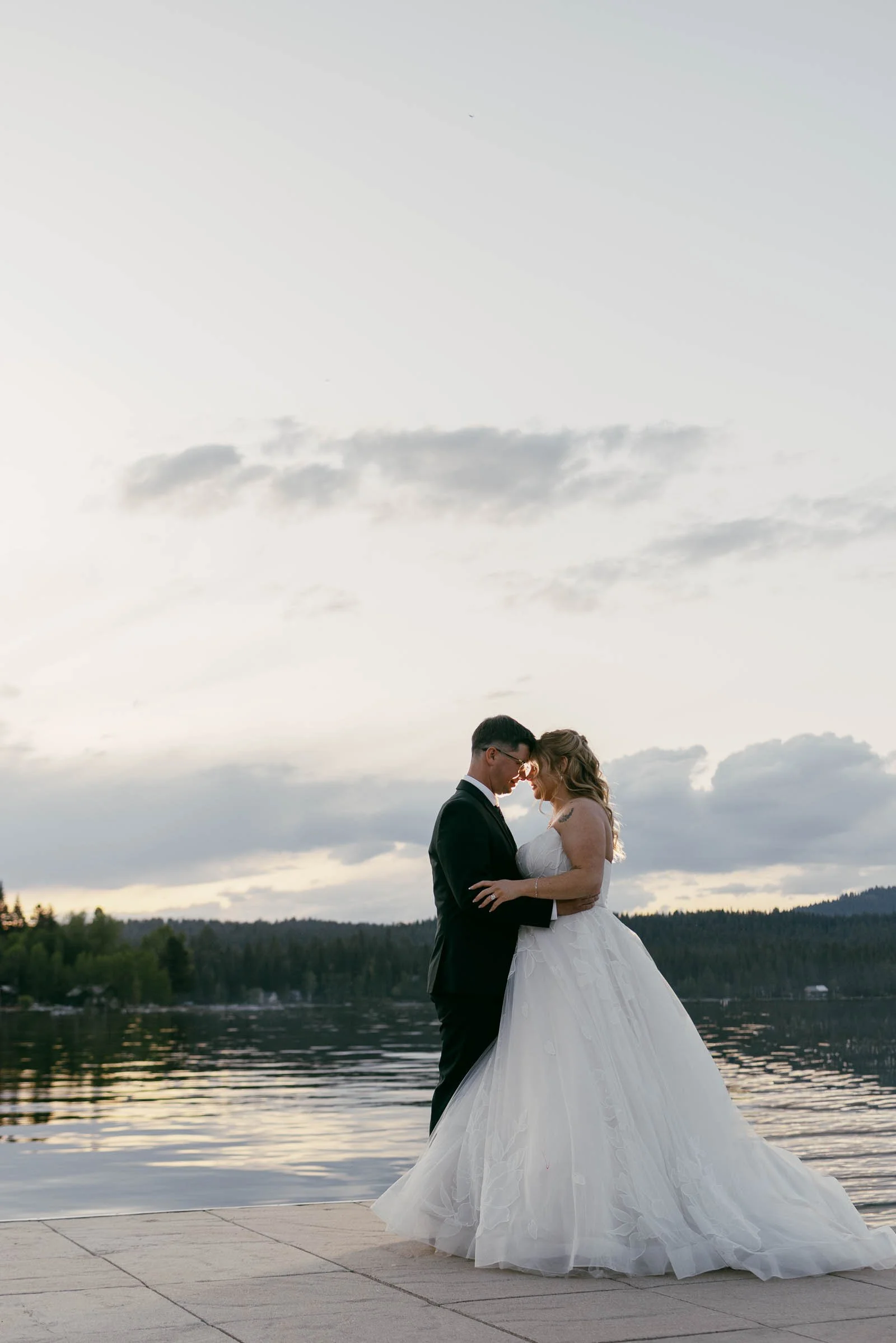  A bride and groom stand close together on a dock by a calm lake at sunset, with trees and mountains in the background and soft clouds in the sky. 