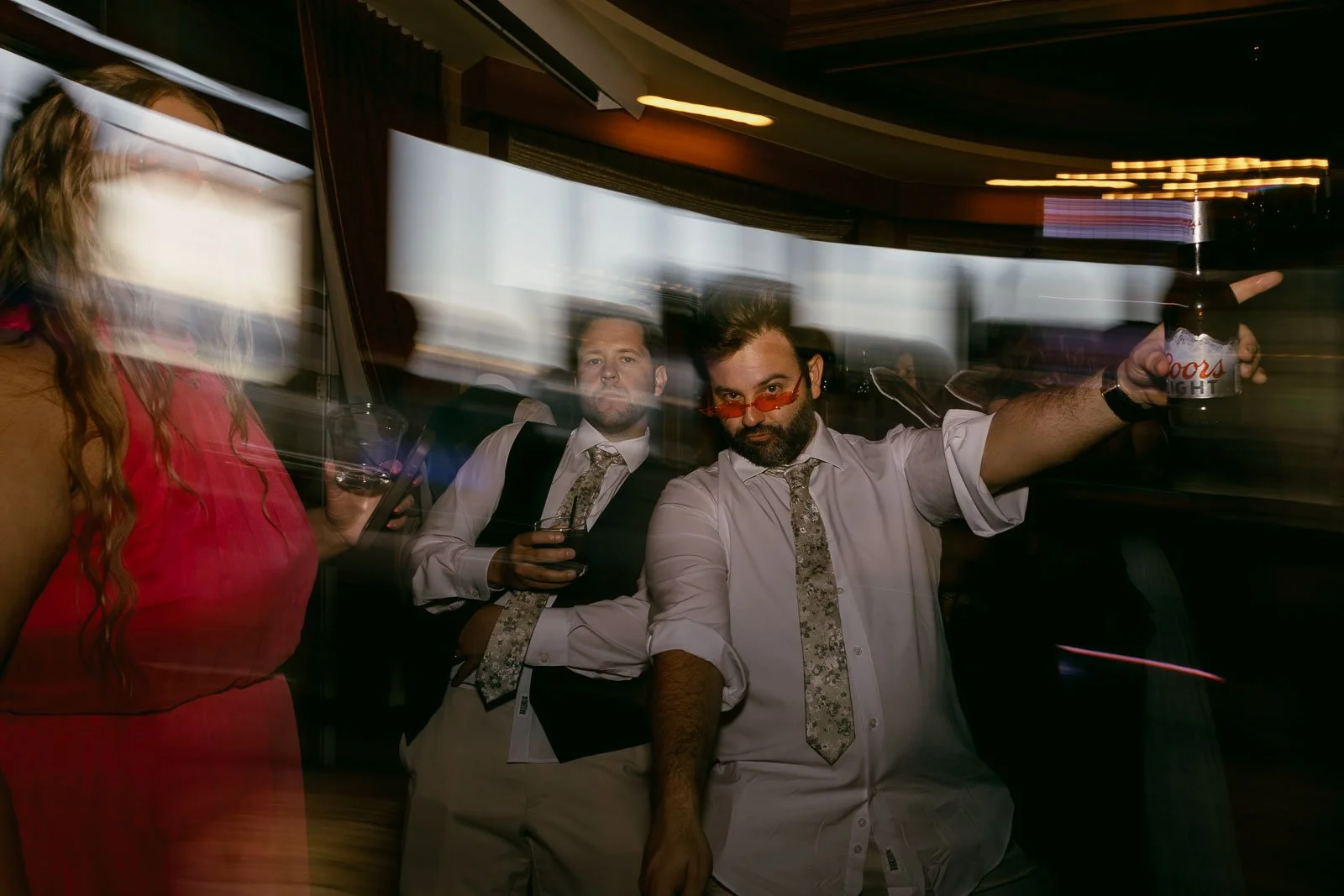  Two men in dress shirts and ties pose on a dance floor with motion blur; one points while holding a drink. A woman in a red dress stands in the foreground, also blurred. The scene appears lively, likely at a party or event. 