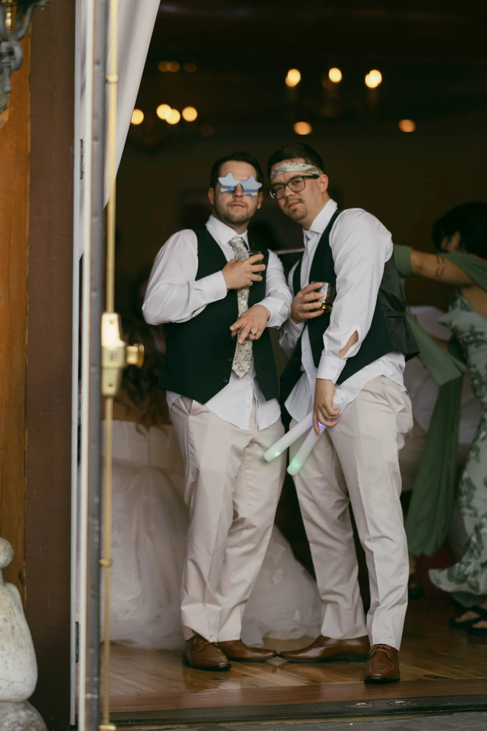  Two men in dress shirts, vests, and sunglasses pose playfully at a party, each holding a drink and a glow stick, with other guests blurred in the background. 