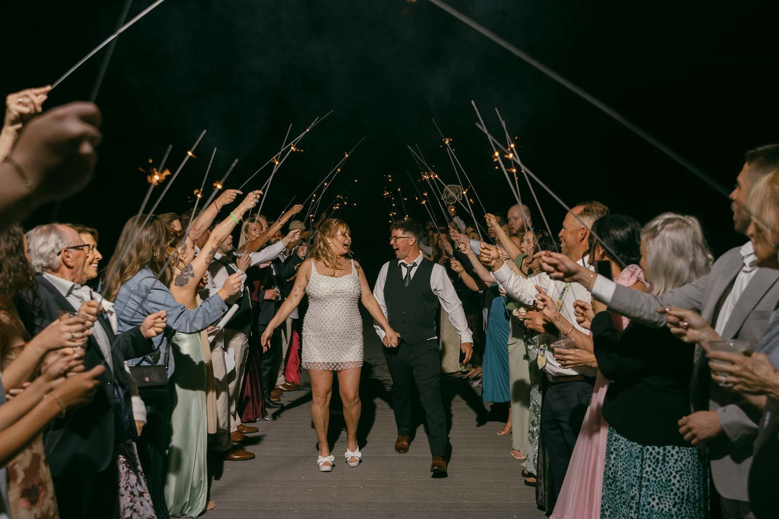  A couple walks joyfully through a crowd of people holding sparklers at night, celebrating. The woman wears a short white dress, and the man is in a vest and tie. Guests cheer and smile on both sides. 