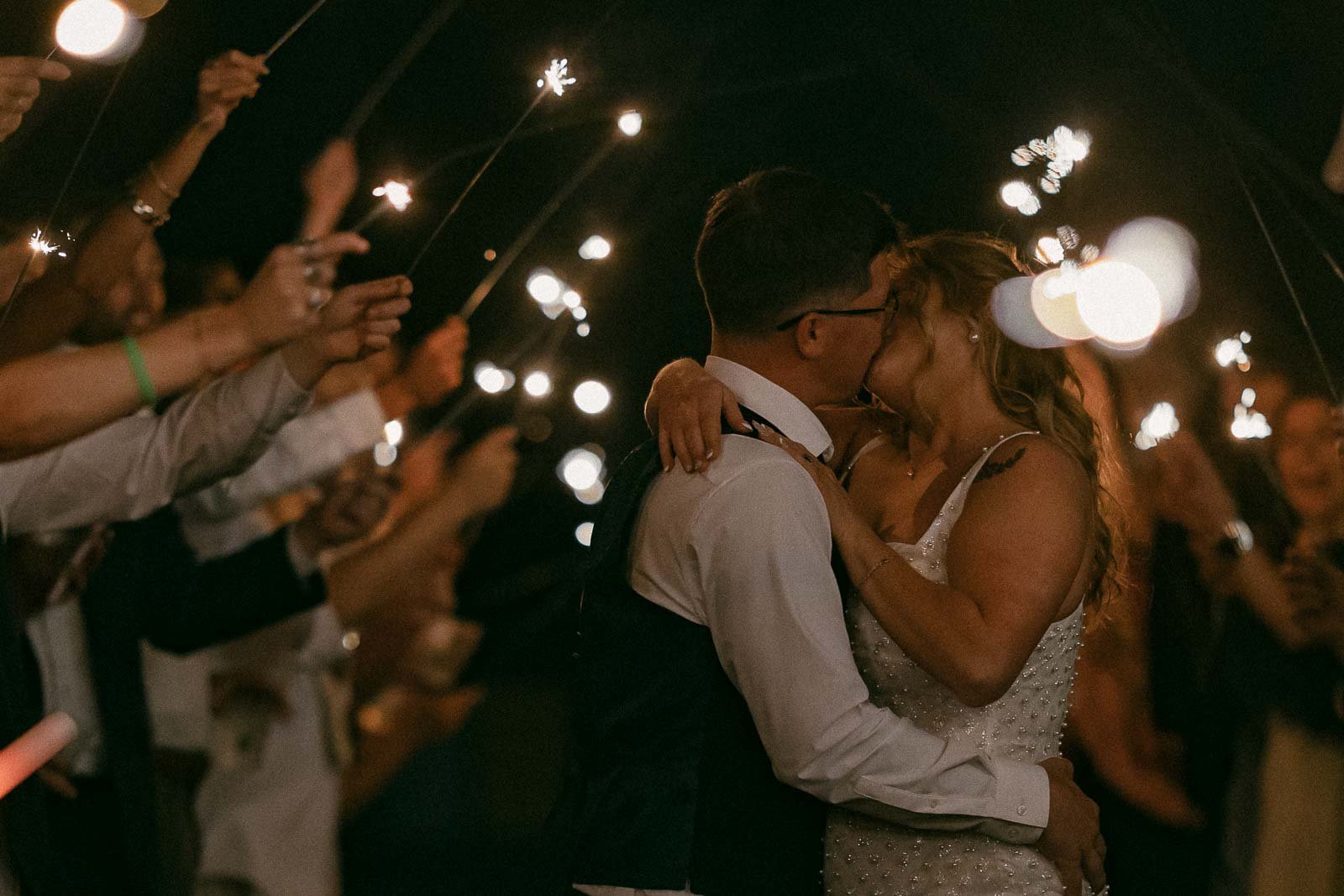  A bride and groom kiss while surrounded by guests holding sparklers at night, creating a festive and romantic atmosphere. 