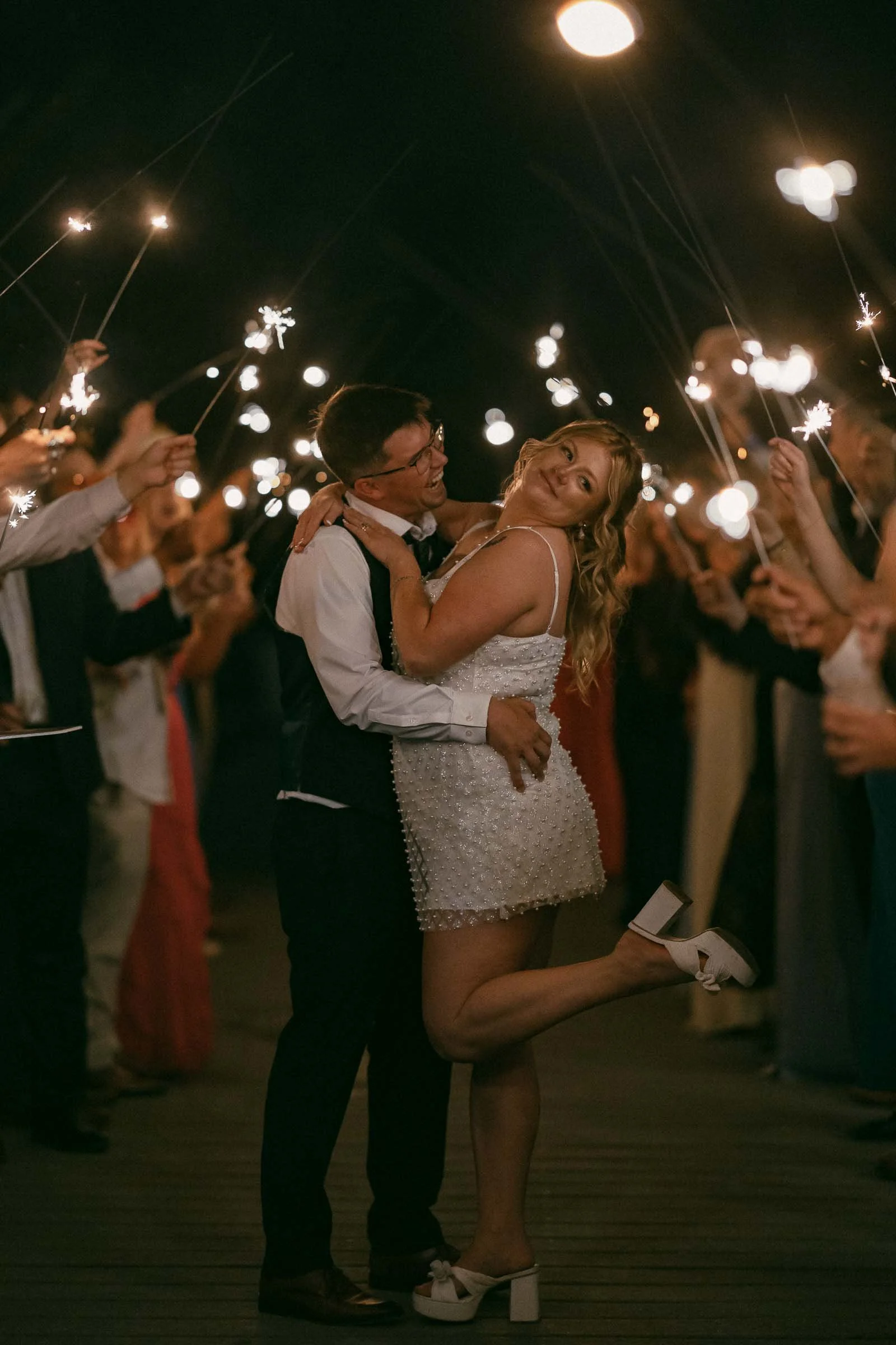  A couple poses happily at night surrounded by people holding sparklers. The woman lifts one leg playfully, smiling, while the man holds her. Everyone is dressed up, creating a festive and joyful atmosphere. 