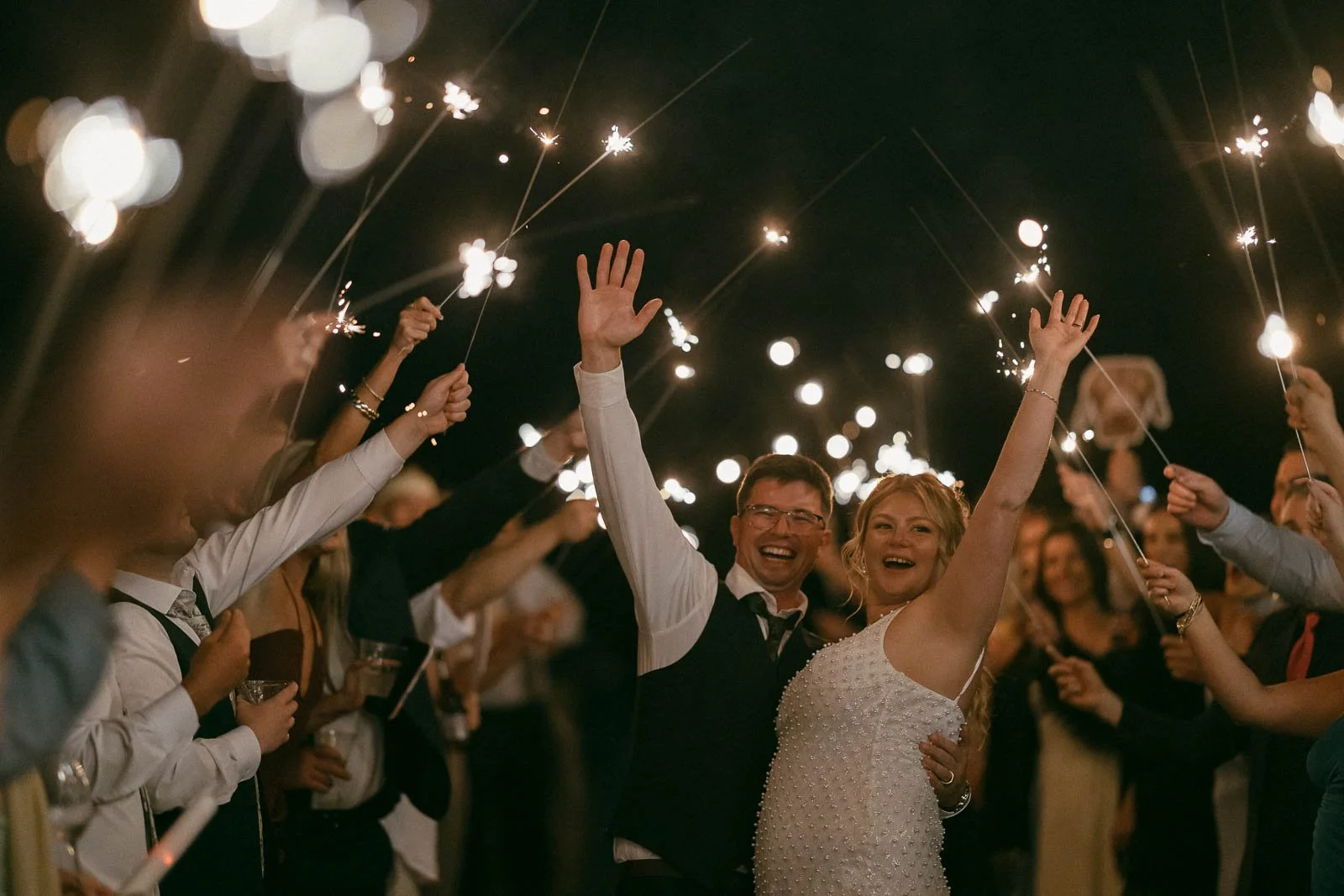 At night, a beaming newlywed couple raises sparklers overhead while walking through a cheering crowd of guests in celebration.
