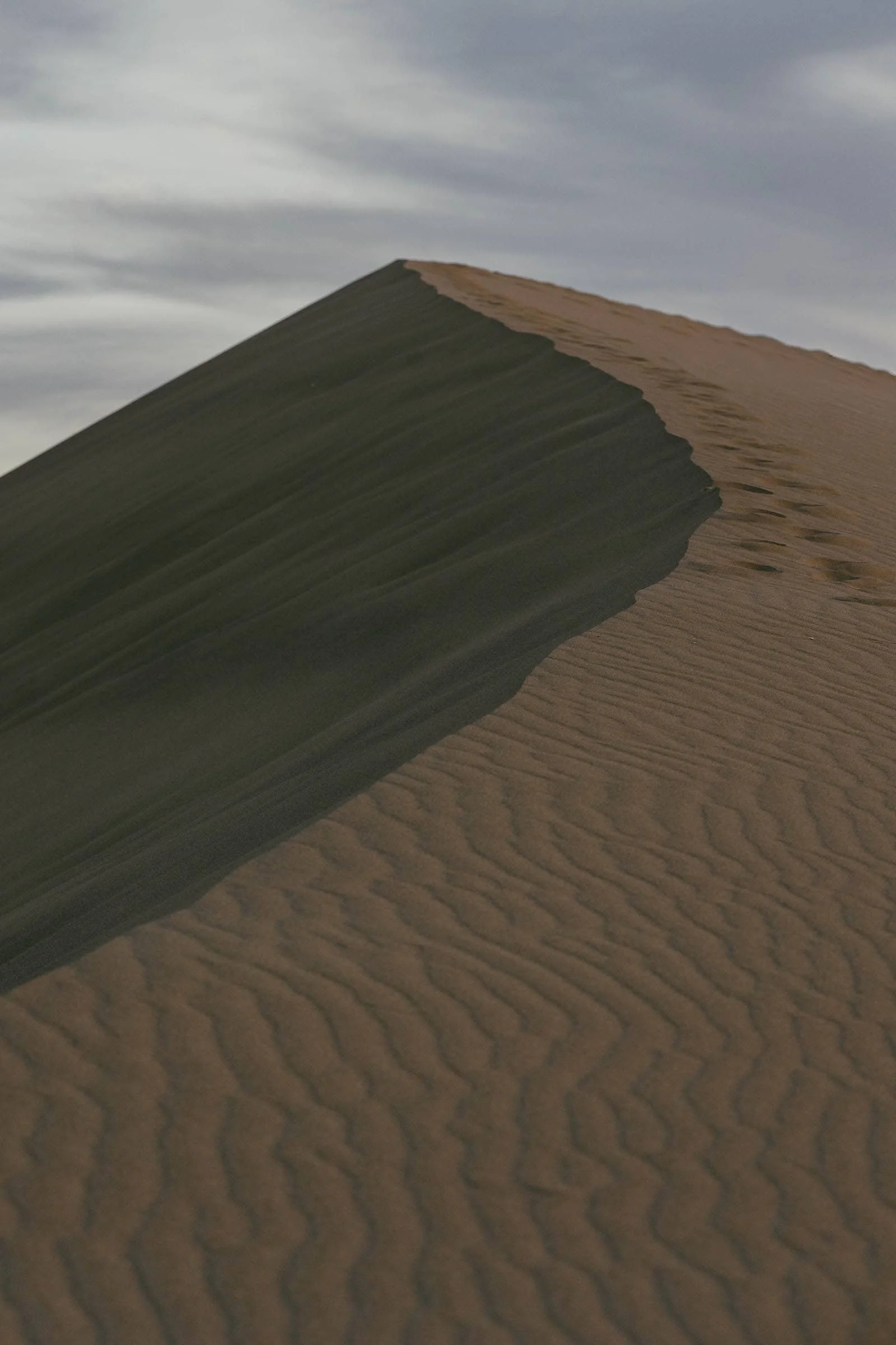  A lone sand dune ridge cuts through the frame, split by a long dark shadow. A trail of footprints runs along the top edge, disappearing into a soft cloudy sky above. 