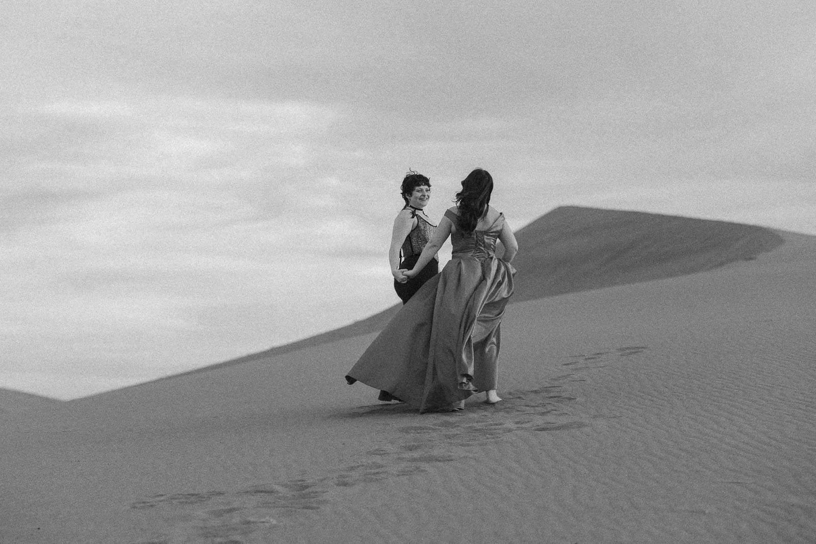  Two women in long dresses walk barefoot through sand dunes, holding hands and smiling. Footprints trail behind them. The scene is in black and white with cloudy skies above. 