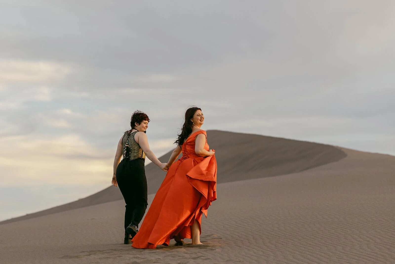  A couple strolls hand in hand across sandy dunes beneath a cloudy sky, glancing back with smiles. One wears a flowing red dress, the other all black, their footprints trailing behind them. 