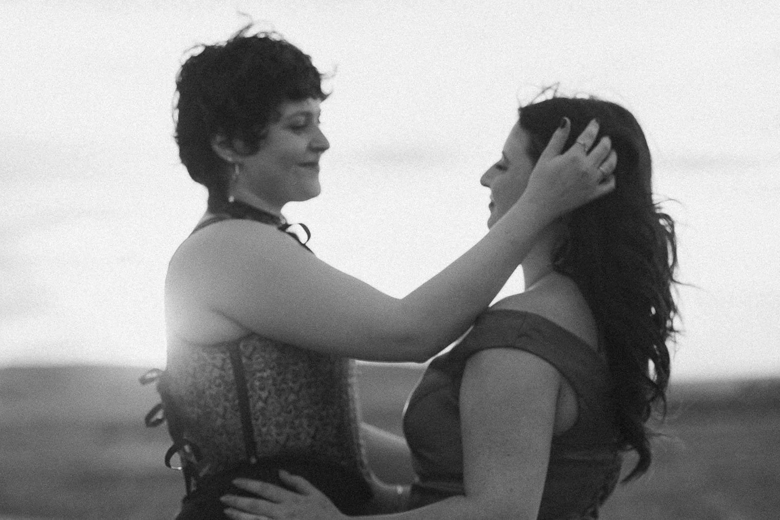  Two people stand close together outdoors, one gently touching the other’s hair. Both appear to be smiling softly, sharing an intimate and affectionate moment. The image is black and white and softly focused. 