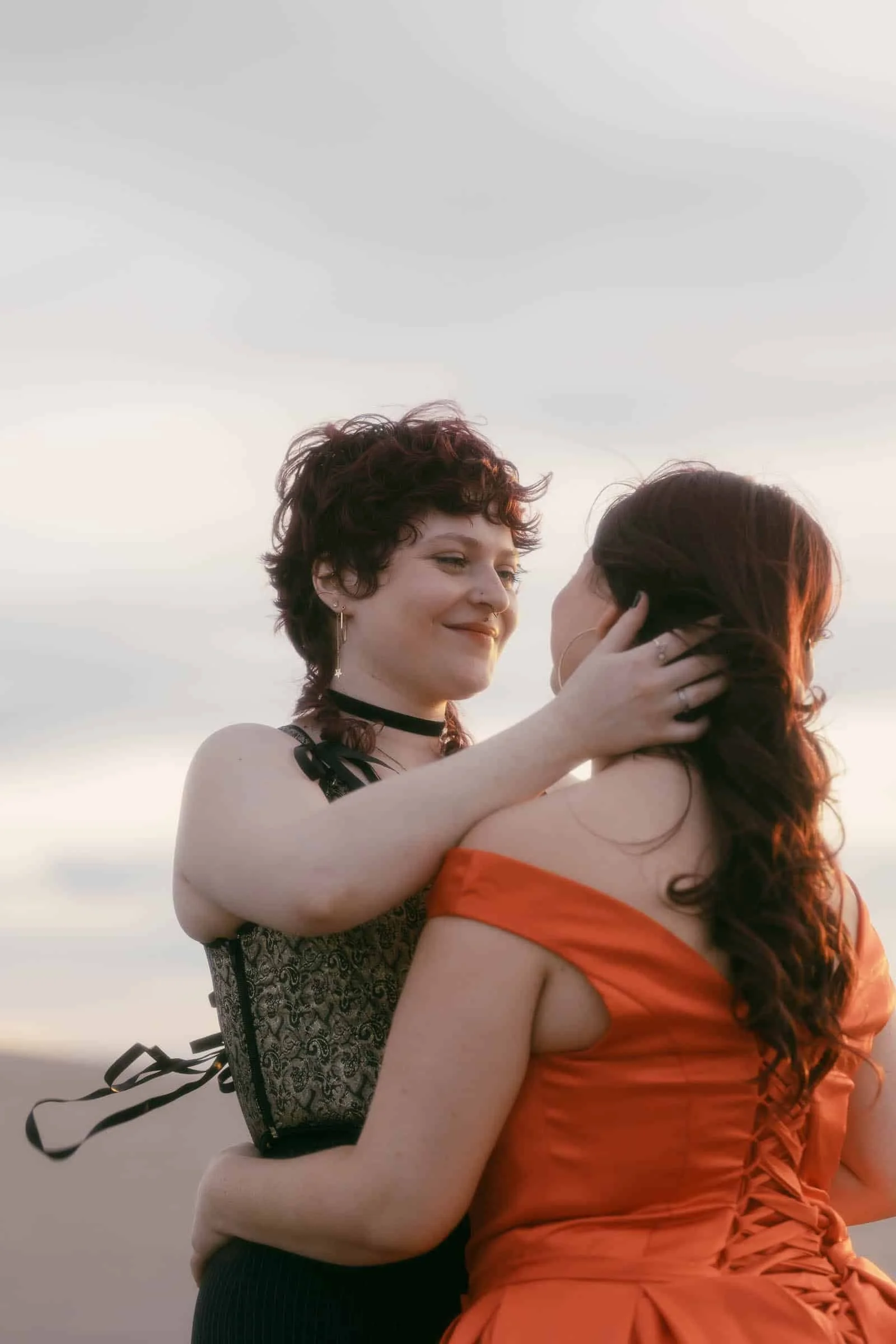  Two women stand close together at sunset, one in a green corset and the other in an orange dress, gently holding each other and sharing an affectionate moment. 