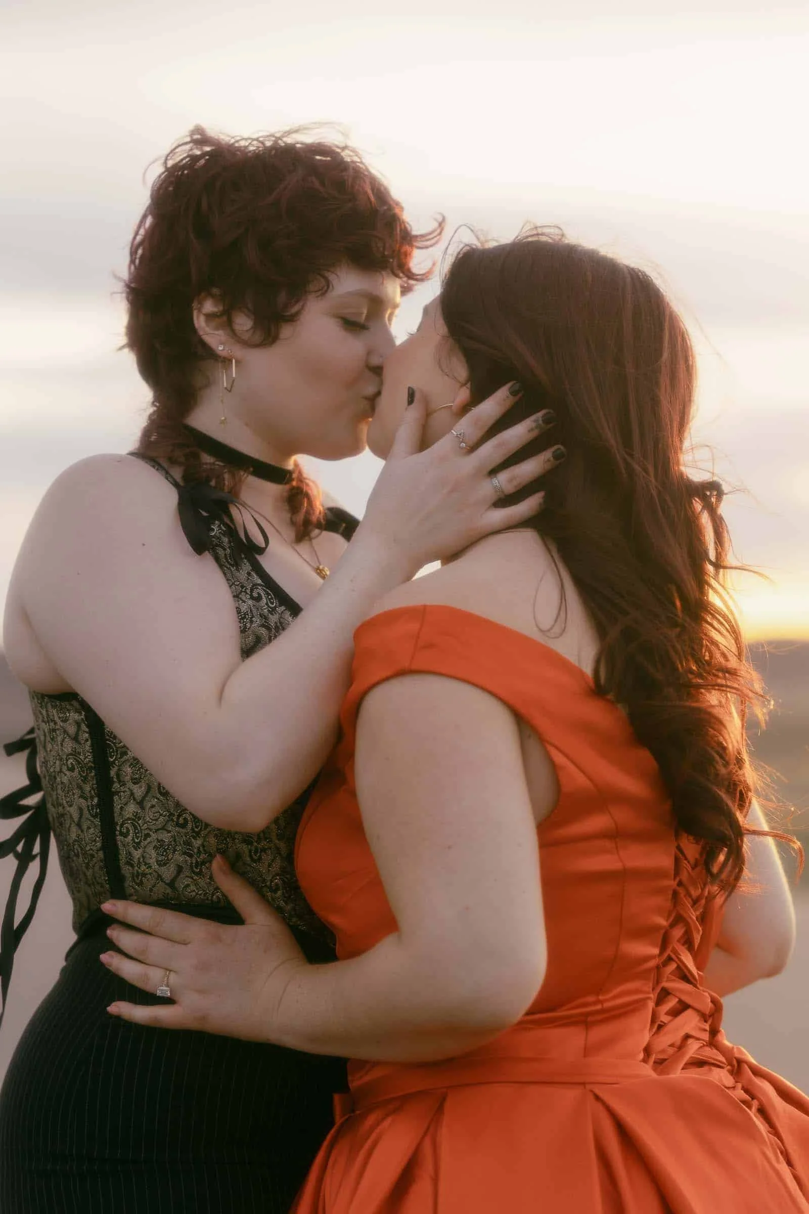  Two women share a kiss at sunset, one in a patterned top and choker, the other in an off-shoulder orange dress. They embrace tenderly, with soft sunlight in the background. 