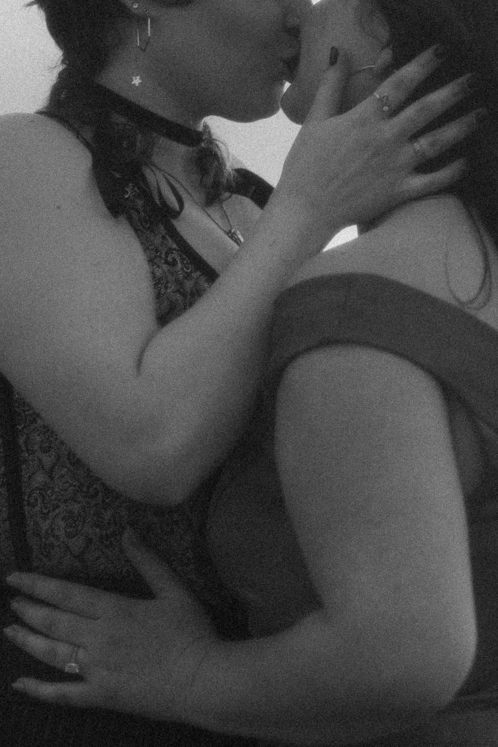  A grayscale close-up of two women sharing a tender kiss, one hand cradling a cheek, the other resting at a partner's back. The image is soft and intimate, shot with a dreamlike focus. 