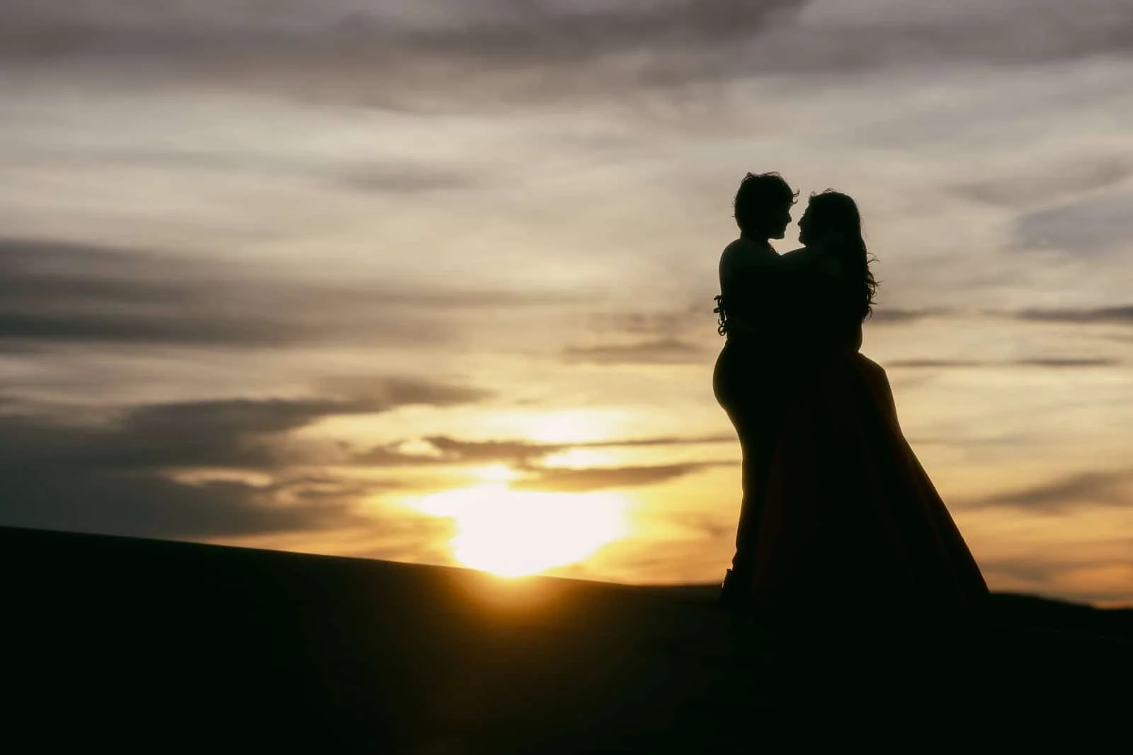  Two people in formal attire embrace, silhouetted against a dramatic sunset sky with clouds, standing on a hill or dune. 