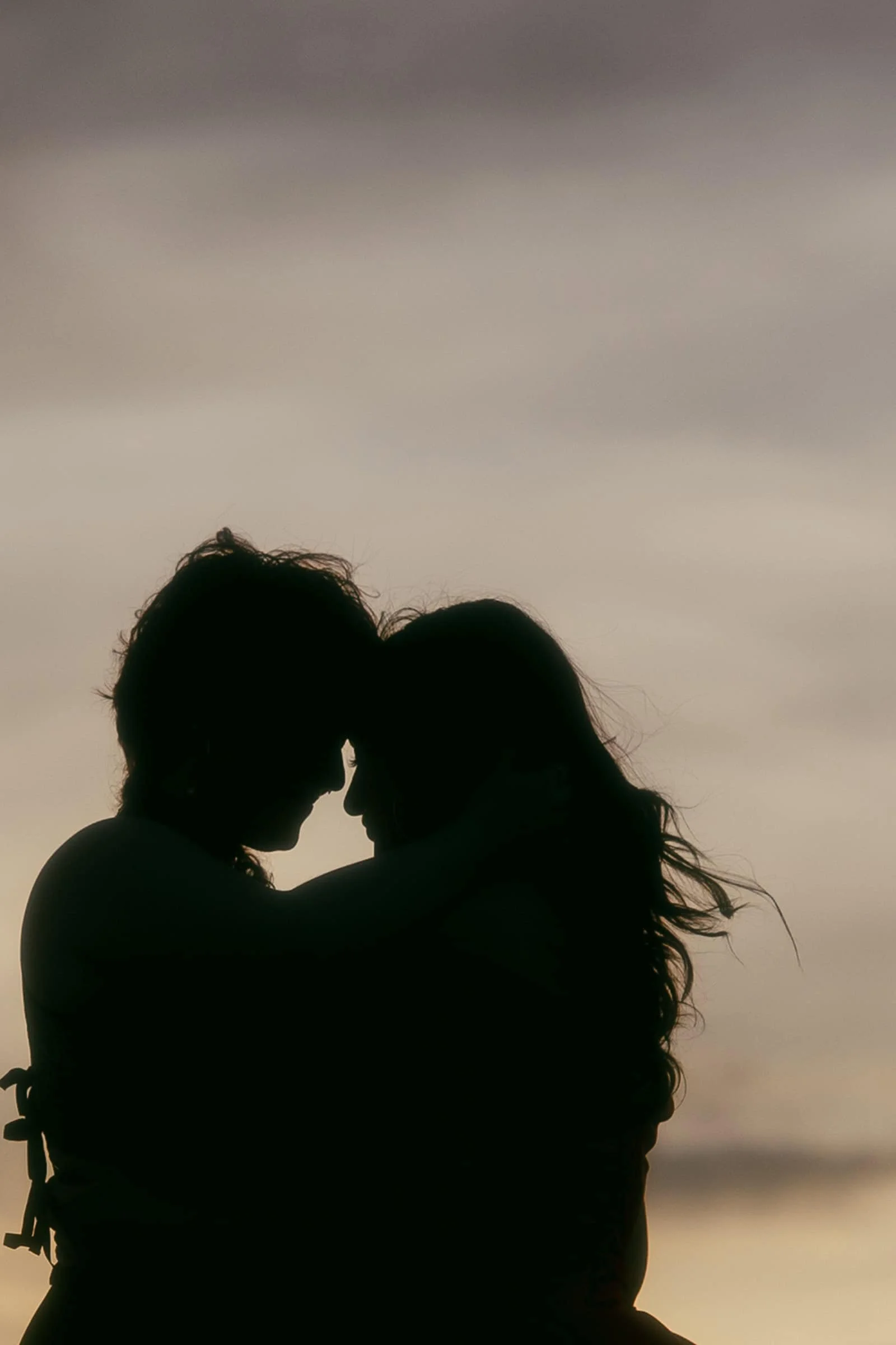  Two figures stand face to face in silhouette, wrapped in each other's presence against a softly lit cloudy sky at sunset. No detail, just shape and feeling. 