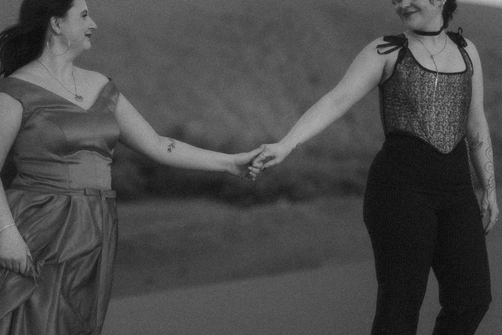 Two people smiling and holding hands, walking together outdoors. One wears a dress, the other wears a sleeveless top and pants. The image is in black and white and slightly blurred. 