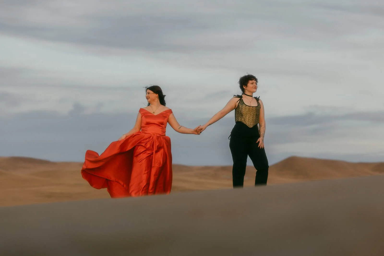  Two people hold hands on a desert landscape. One wears a flowing red dress, the other wears black pants and a gold top. Both look in opposite directions under a cloudy sky. 