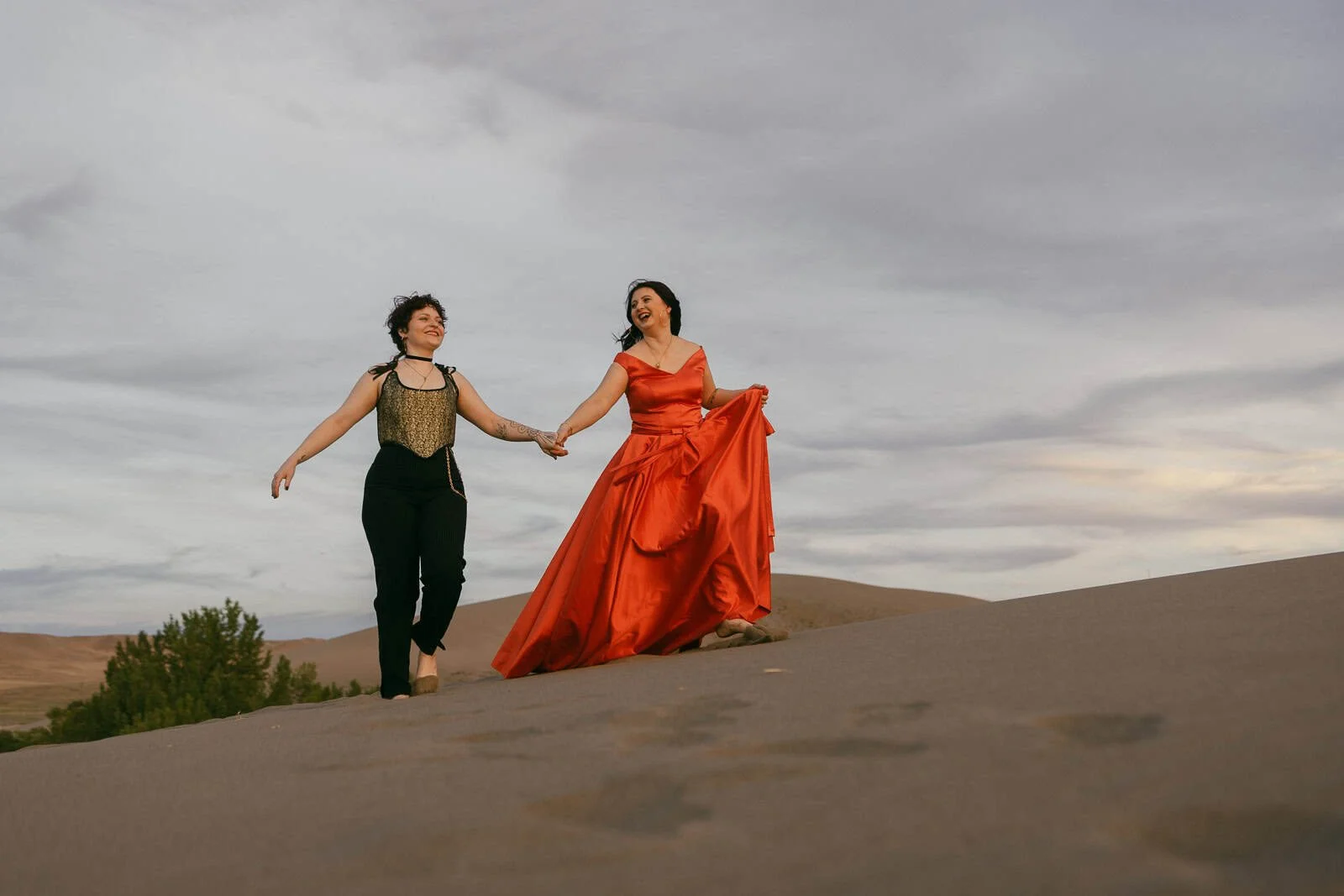  Two people holding hands walk on sand dunes; one wears a gold top with black pants, the other a flowing red dress. Both are barefoot, smiling, and surrounded by a cloudy sky and distant trees. 