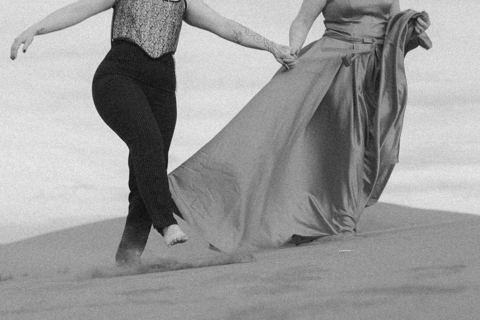  A black and white image of two women walking across open sand, fingers intertwined. One wears a corset and pants, the other a long flowing dress trailing behind her. 