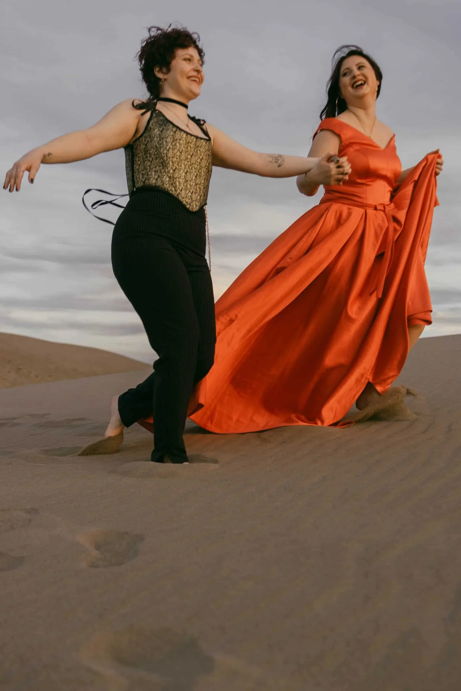  Two women laugh and dance on sand dunes at dusk. One wears black pants and a lace top; the other wears a flowing red gown that billows in the wind as they hold hands. 