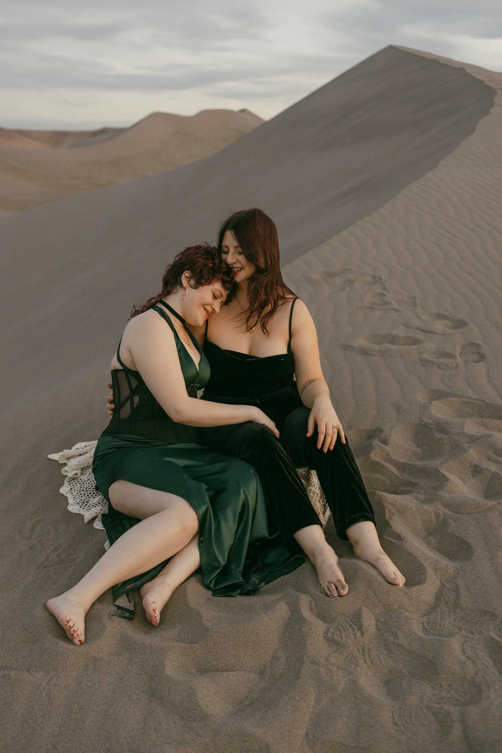  Two women in dark dresses sit side by side on the dunes, bare feet sinking into sand, laughing and leaning into each other. Footprints and rolling dunes stretch out peacefully behind them. 