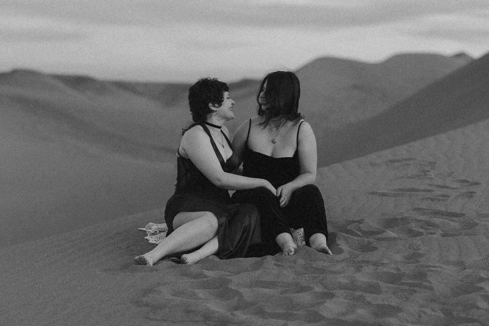  Two people in dresses sit barefoot on sand dunes, facing each other and smiling, with one hand touching. The background shows rolling sand dunes and a cloudy sky in black and white. 