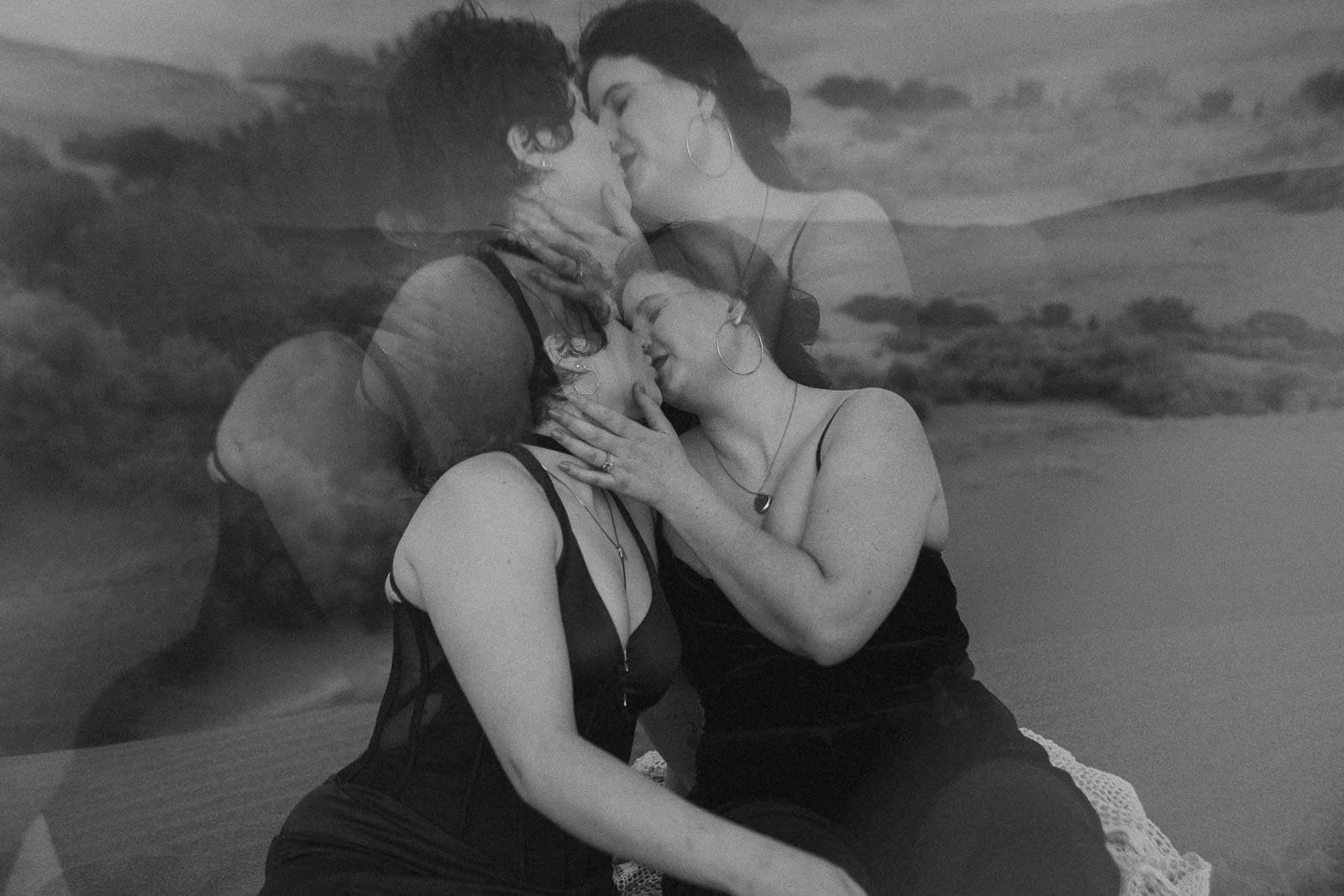 Two women in black clothing share an intimate moment, embracing and touching faces, with a double exposure effect blending their images against a natural, outdoor background. 