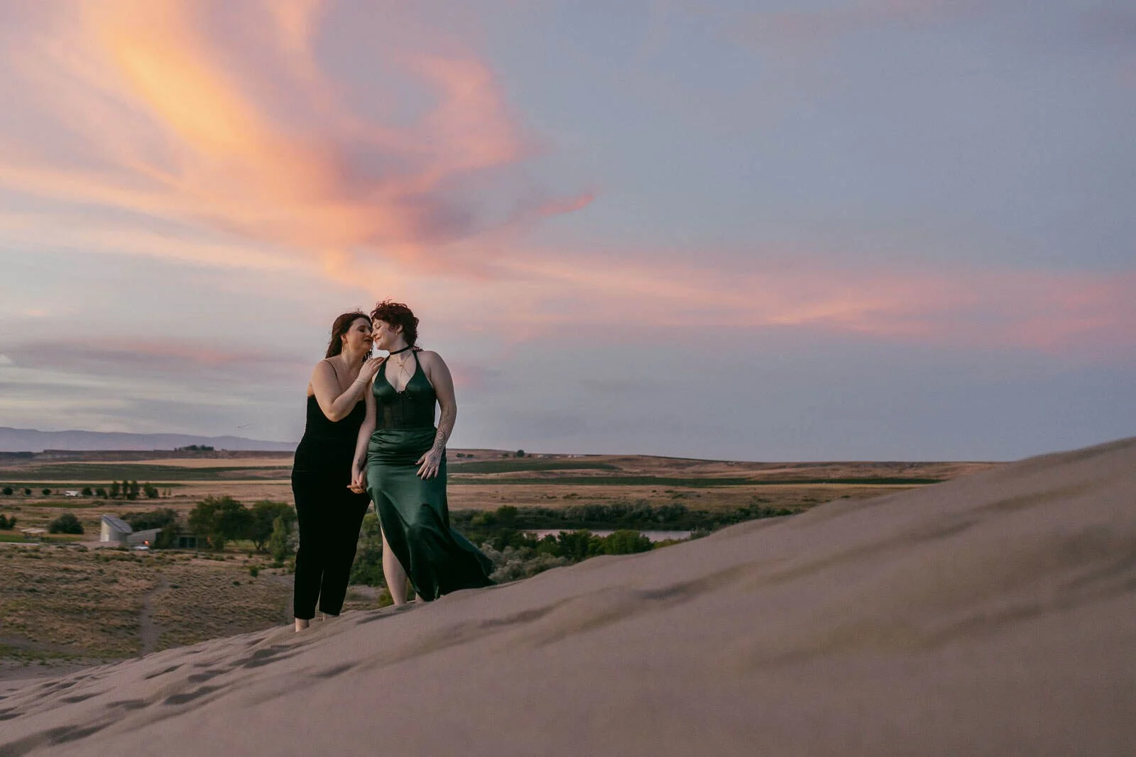  Two people stand close together on a sand dune at sunset, sharing a tender moment. The sky is painted with pink and orange clouds, and open landscape stretches out in the background. 