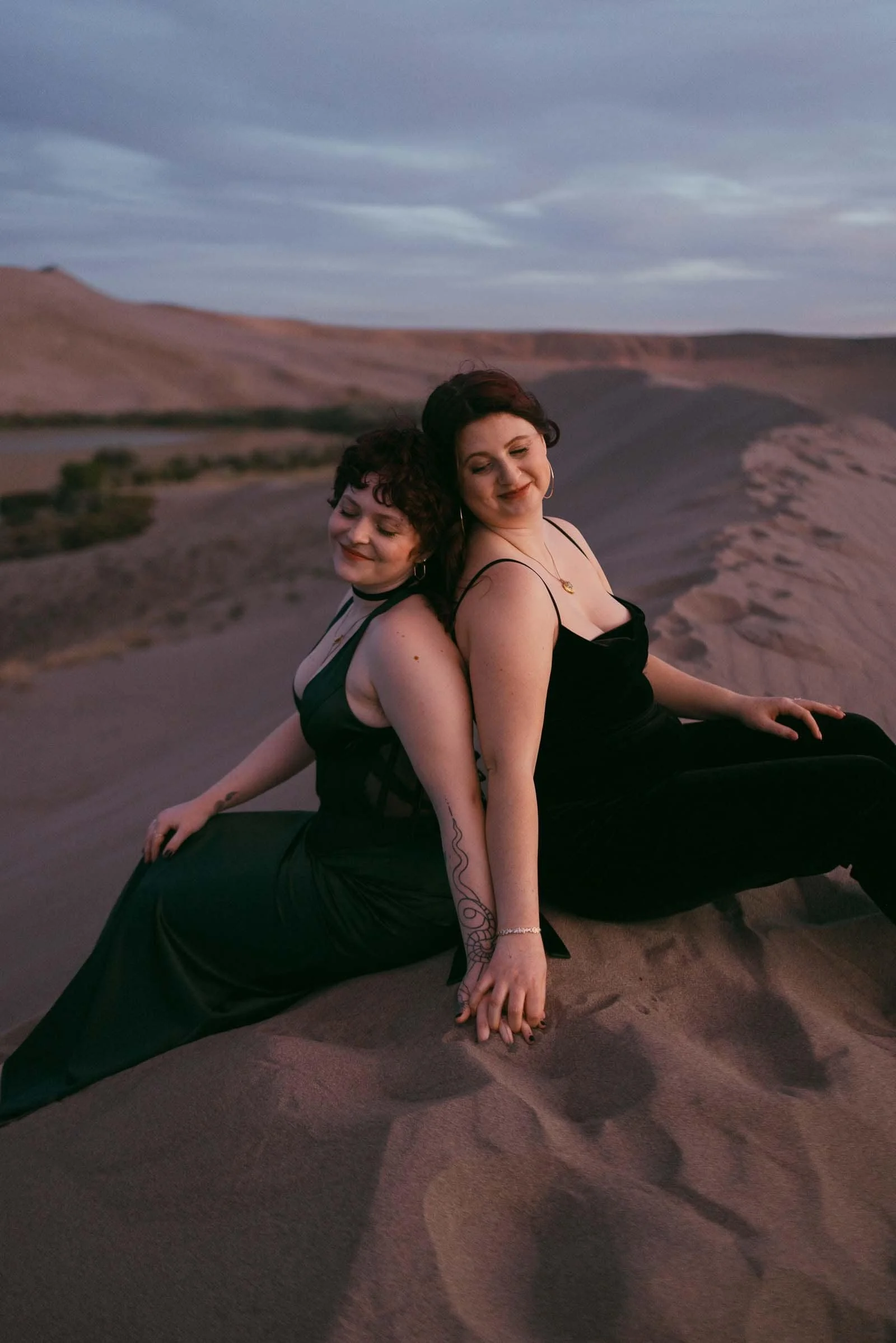  Two women in black dresses sit back-to-back on sandy dunes at sunset, holding hands and smiling softly with closed eyes, creating a peaceful and intimate scene in a natural desert landscape. 