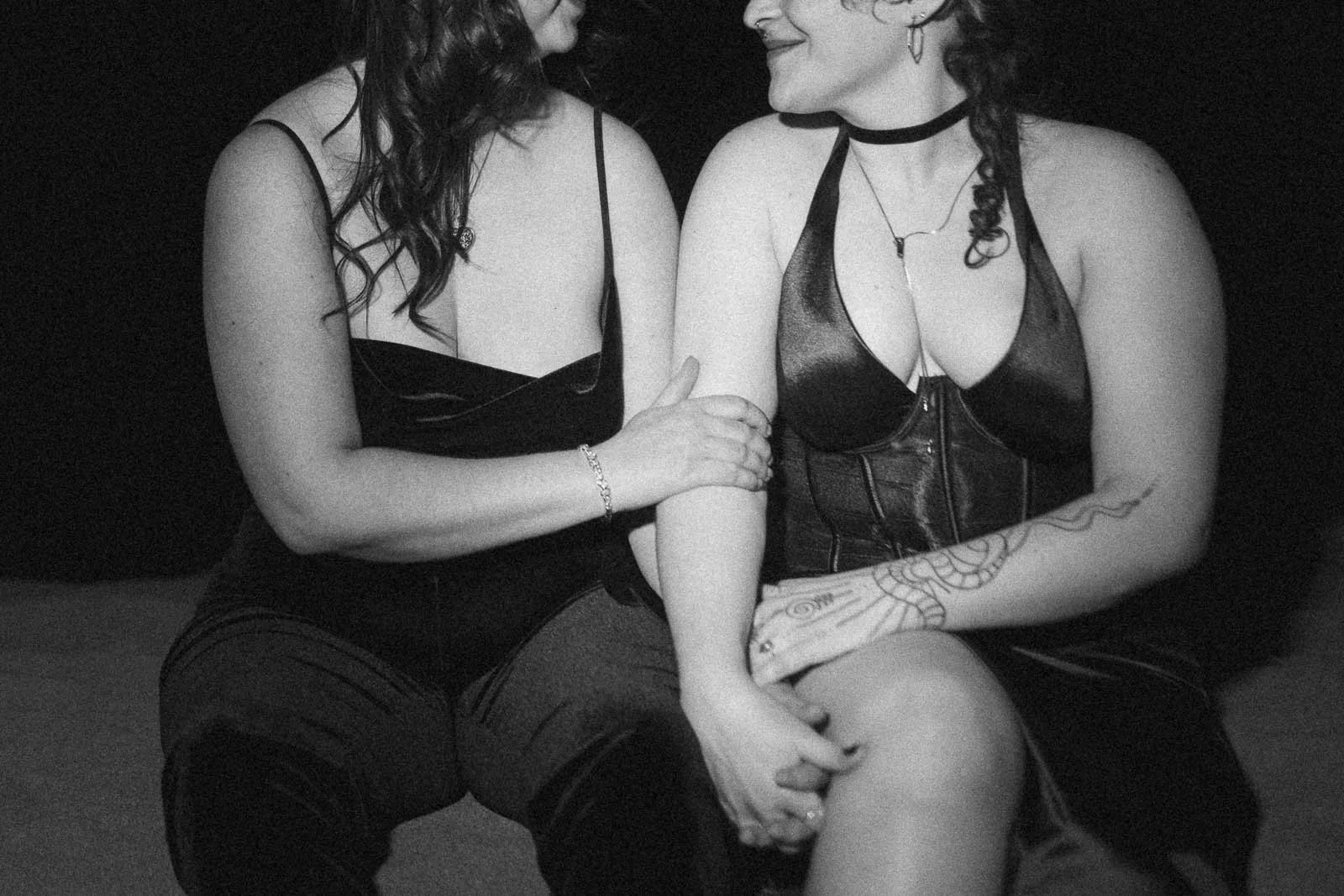  Two people sit close together, smiling at each other. They are both wearing dark sleeveless outfits and jewelry. One has a visible arm tattoo and choker necklace. The image is black and white. 