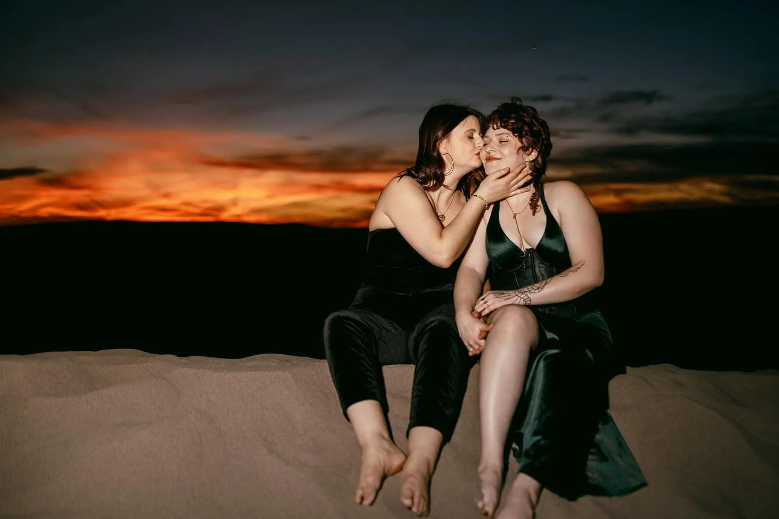At sunset on the sand, one woman presses a soft kiss to her partner's cheek. Both wear dark dresses, hold hands, and smile as a warm orange and pink sky glows behind them.