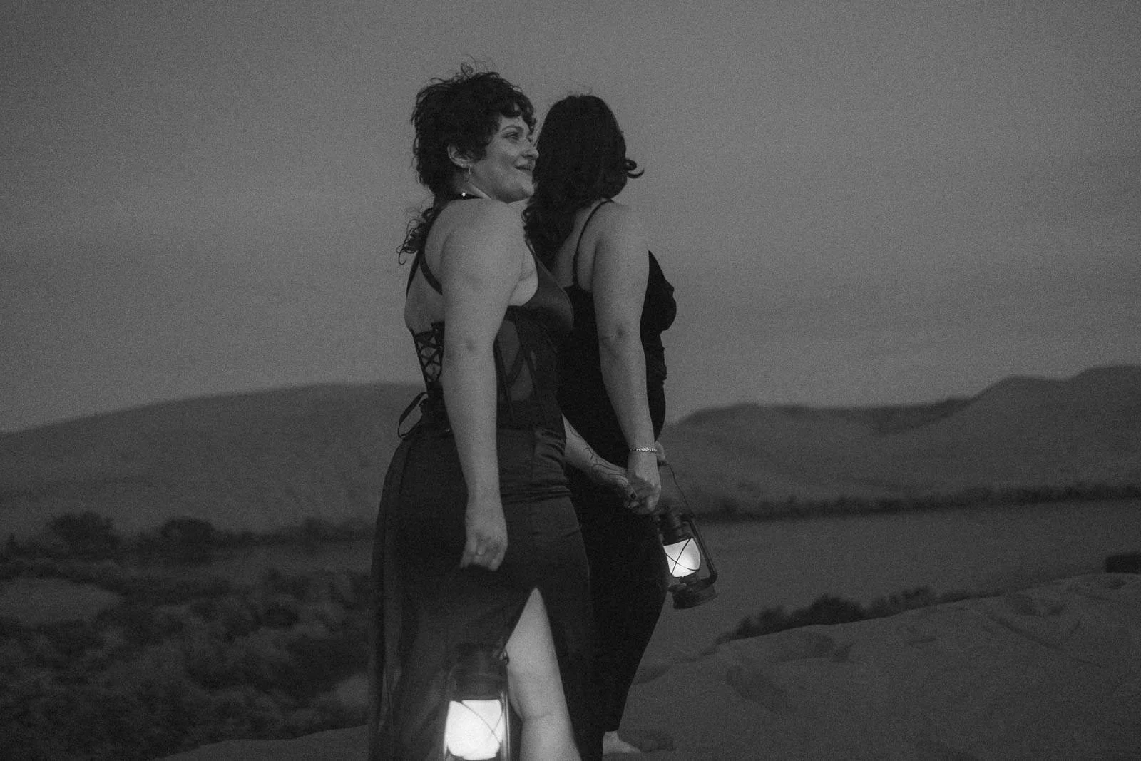  Two people in black dresses hold lanterns and walk on a sandy hill at dusk, with blurred hills and water in the background. The image is in black and white. 