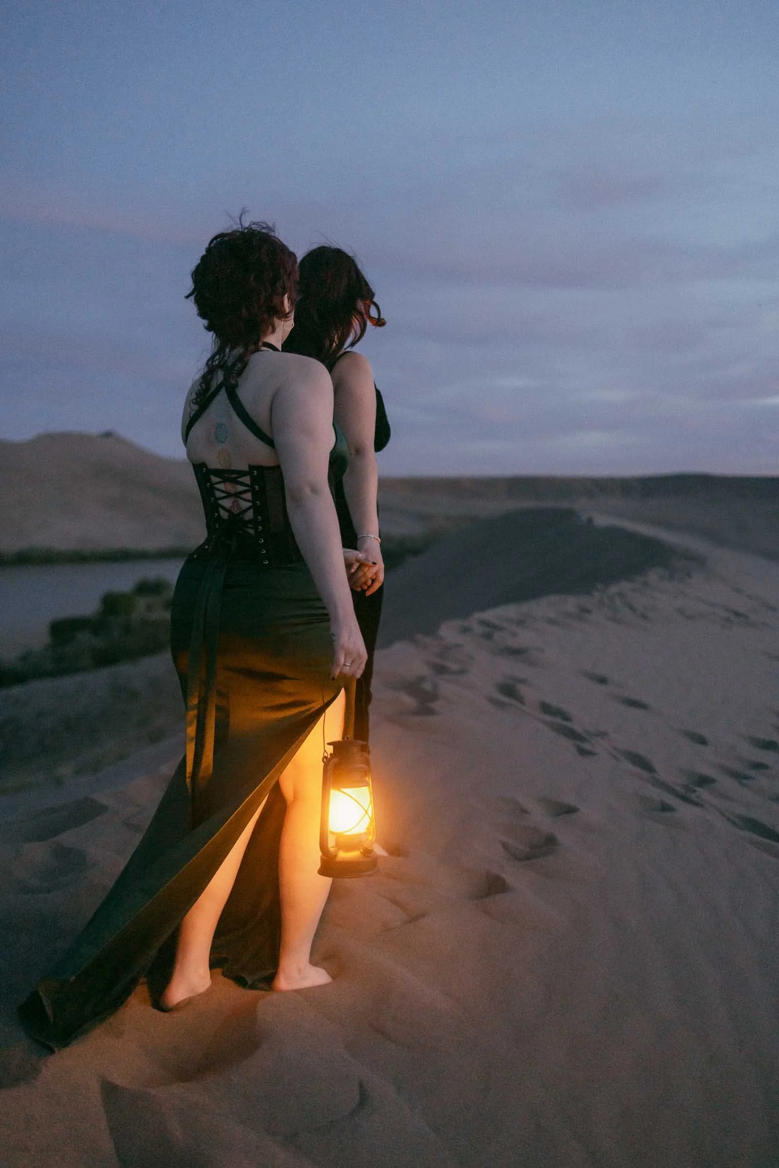  Two people in long black dresses stand barefoot on sand dunes at dusk, holding hands. One holds a glowing lantern, illuminating the sand as they look toward the horizon under a blue and purple sky. 