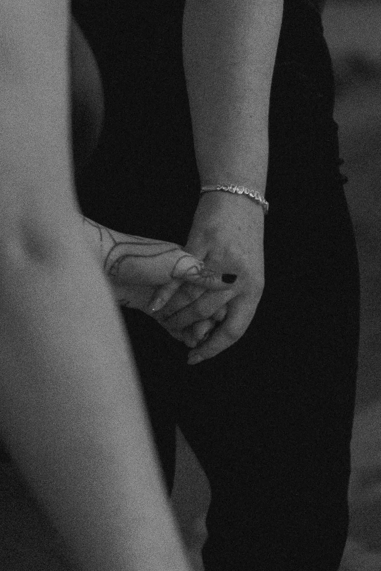  Two people are holding hands, one wearing a bracelet and the other with a visible tattoo. The image is in black and white with a soft focus on their hands. 