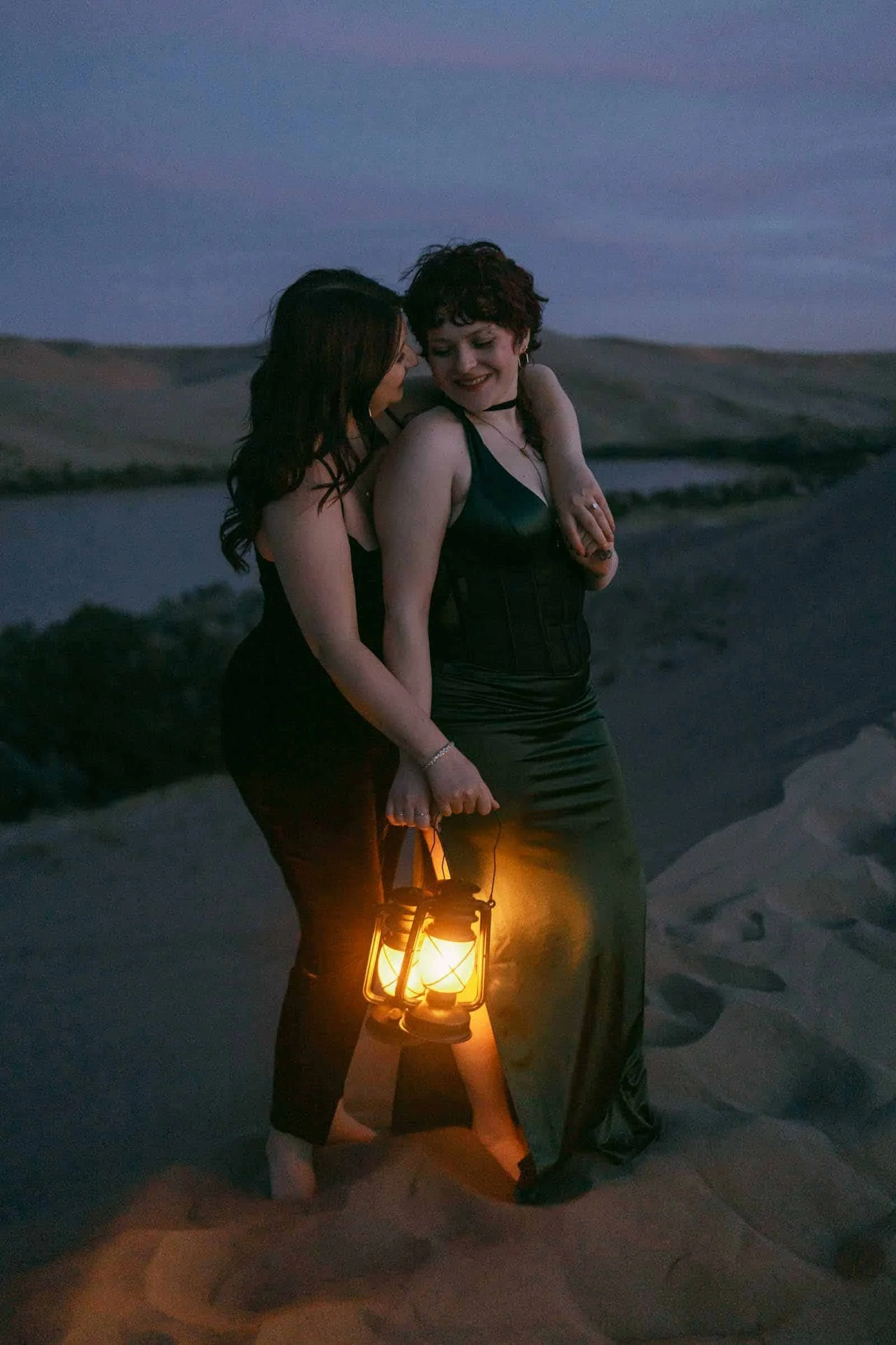 Two women share a close moment at dusk on the desert floor, one holding a softly glowing lantern. They smile toward each other as the Snake River plain and distant hills fade into the evening behind them. 