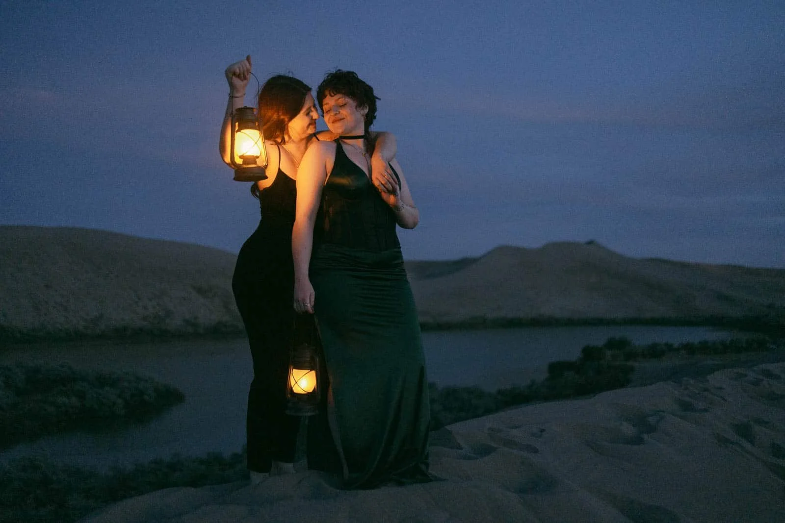 Two women stand together on the dunes at blue hour, each cradling a glowing lantern. One rests her head on the other's shoulder as the dim desert sky and distant hills stretch behind them.