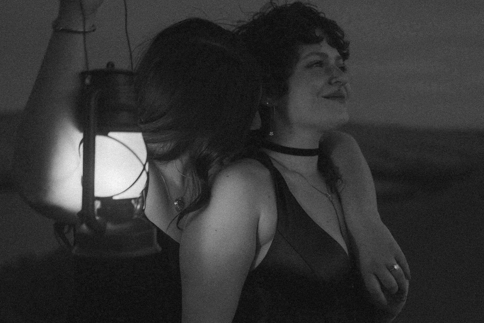A black and white nighttime portrait of two women standing close, one arm wrapped around the other's shoulder, a lantern casting soft light between them. Dark dresses, layered necklaces, and a quiet intimacy that feels like the end of a perfect night